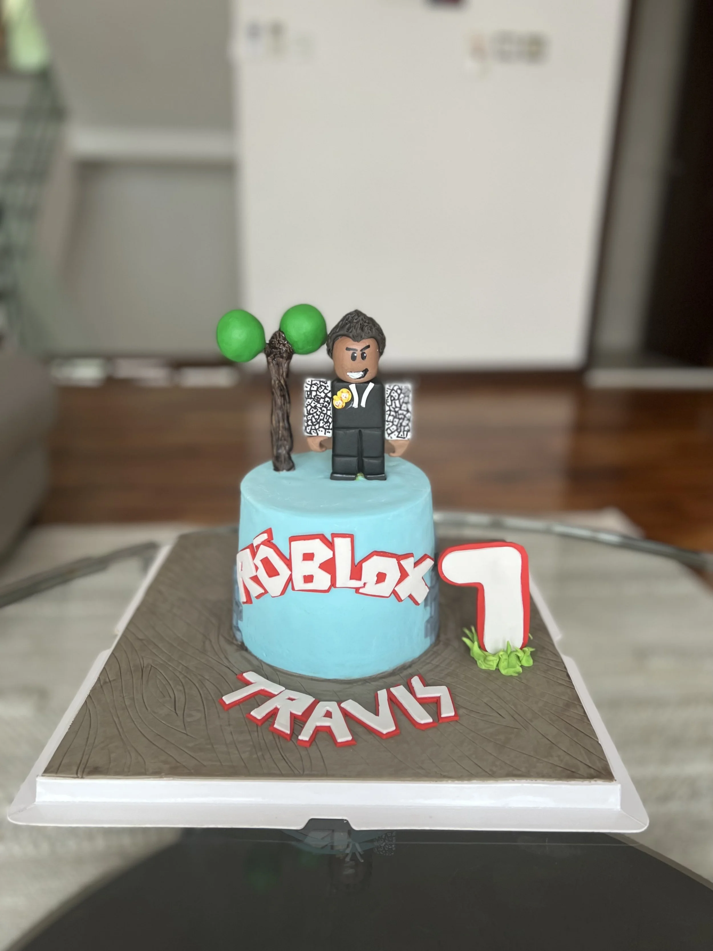 Roblox-themed birthday cake with a fun character figure on top, green trees, a large plane number 7, and the words 'Roblox' and 'TRAVIS' on the cake base.