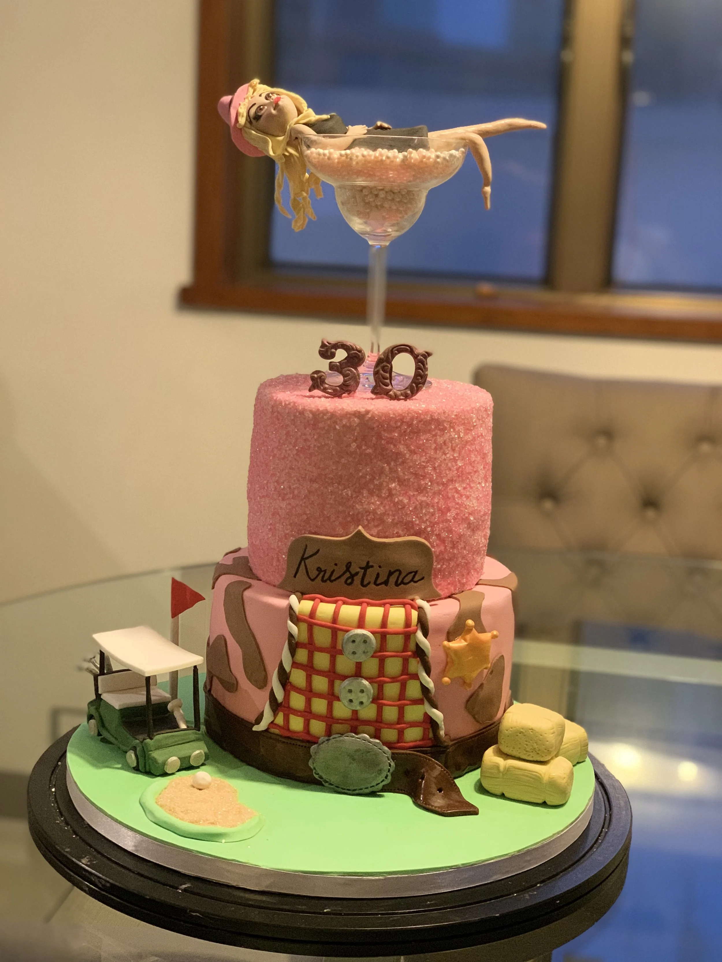 A themed birthday cake decorated with a golf course, golf caddy, golf bag, and golf balls, with two tiers, pink and green, displaying the name Kristina. Toppers include a miniature golf scene on top, a 30th birthday sign, and a doll figurine lying in