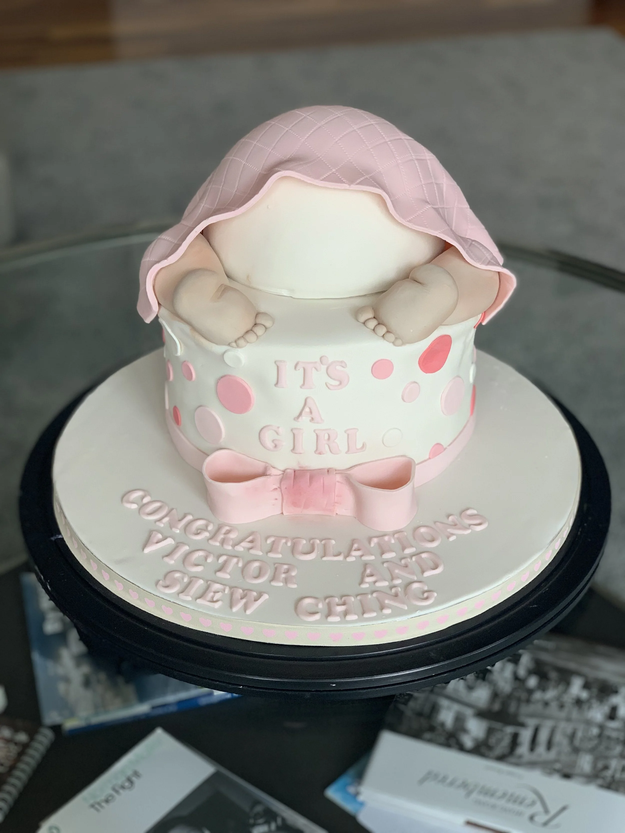 Baby girl-themed cake with a pink quilted blanket draping over a fondant baby figure the back of the cake displays the words 'It's A Girl' with pink and red polka dots, a pink bow, and the message 'Congratulations Victor and Siew Ching' on the base.