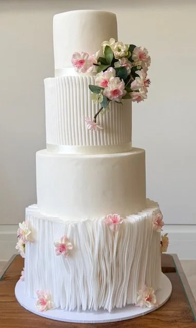 Three-tier white wedding cake with pink and white flowers and vertical and textured patterns.