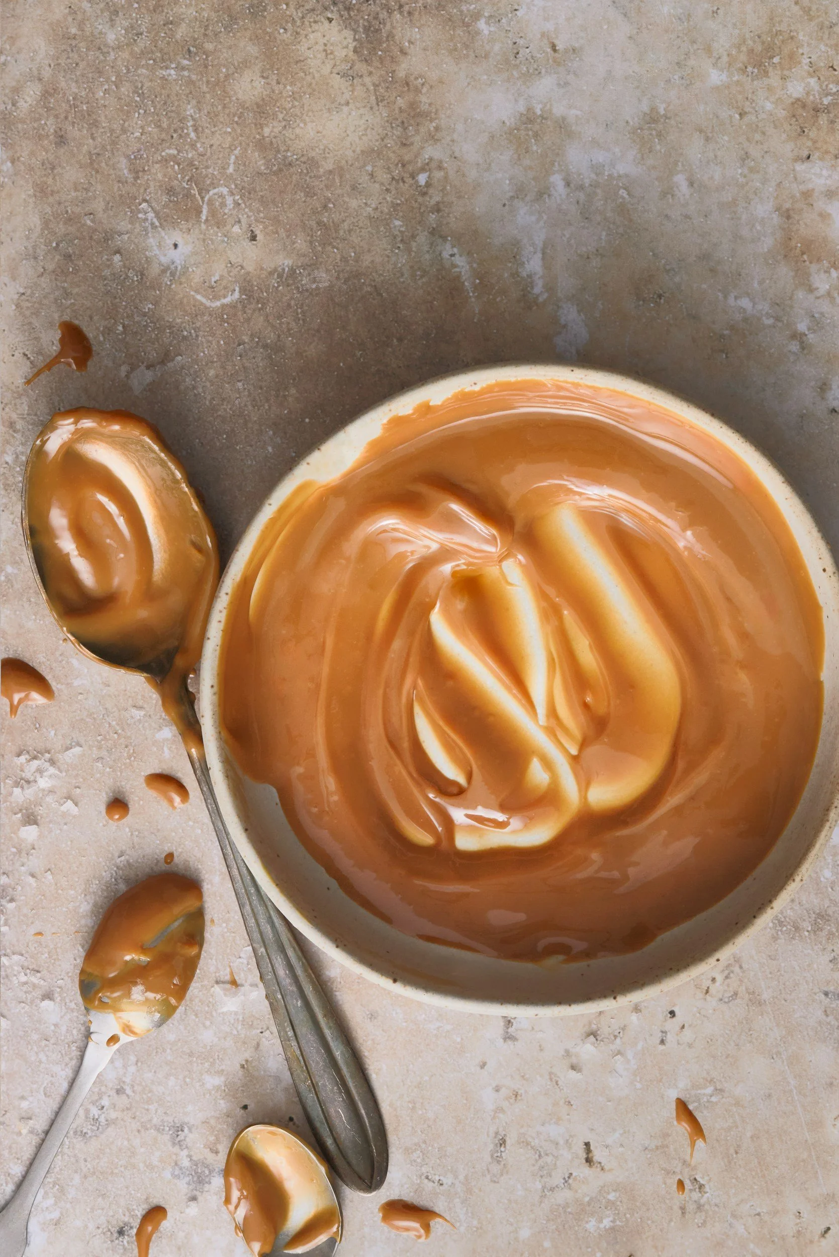 A bowl of creamy peanut butter with a spoonful resting on the side, some spilled around on a textured surface.