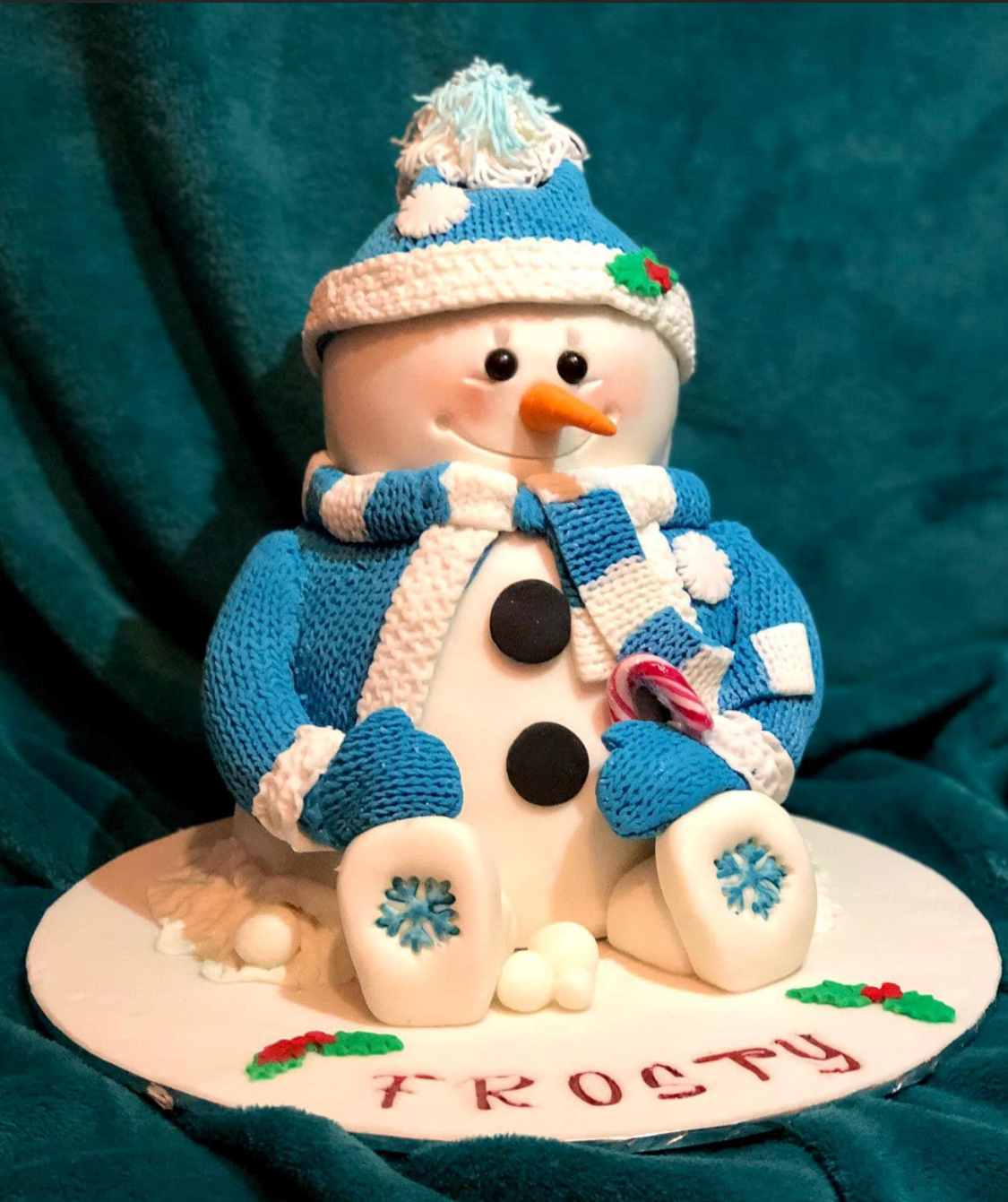 A snowman figurine dressed in a blue and white knit hat and matching scarf, with a carrot nose, black eyes, and black buttons on its body, sitting on a decorative base with the word 'FROSTY' and holly leaves. The snowman is holding a red and white st