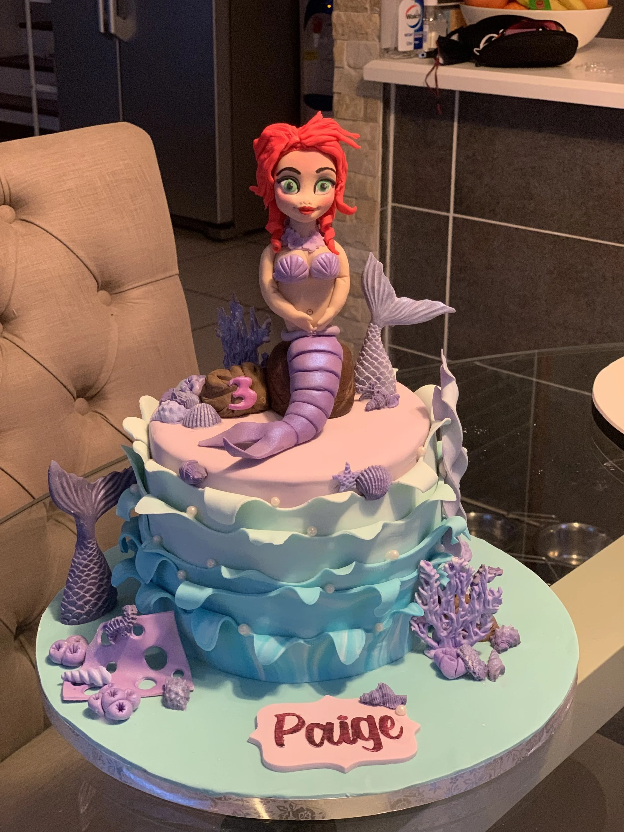A mermaid-themed birthday cake with a figurine of Ariel, the mermaid, sitting on top. The cake is decorated with seashells, coral, and fish decorations in pastel colors. A pink sign with the name "Paige" is attached to the cake.