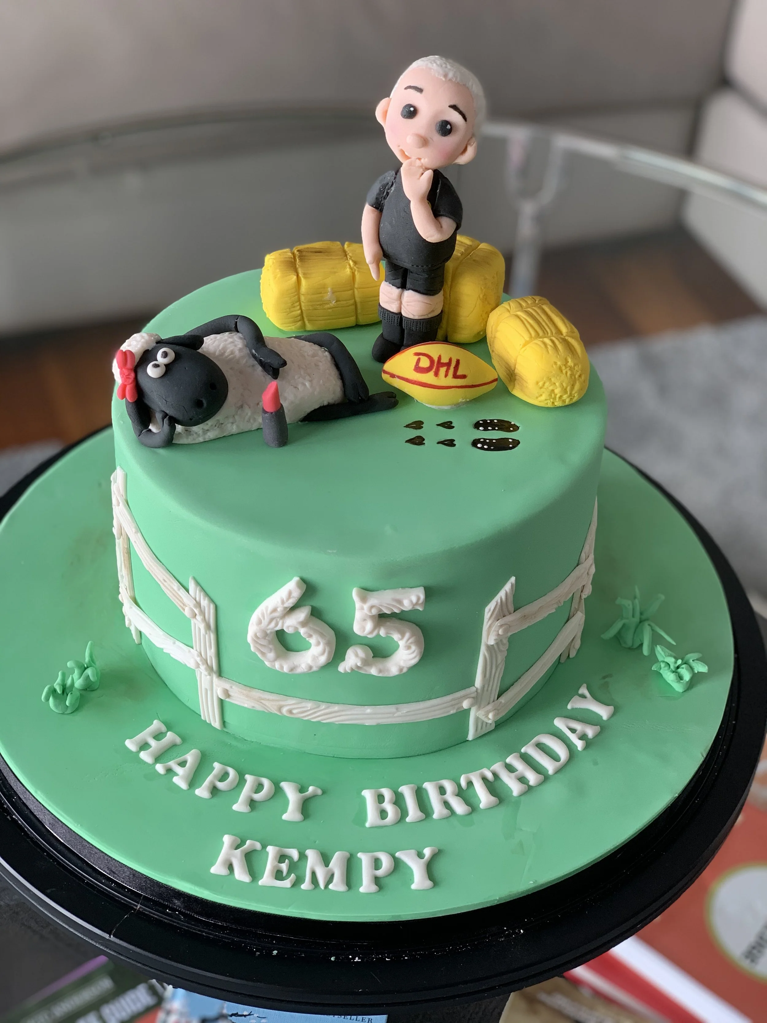 A green birthday cake with a rugby player figurine, a dog figure, a DHL rugby ball, and yellow Tackle dummies on top. The cake has the number 65 and the message "HAPPY BIRTHDAY KEMPY" written on it.