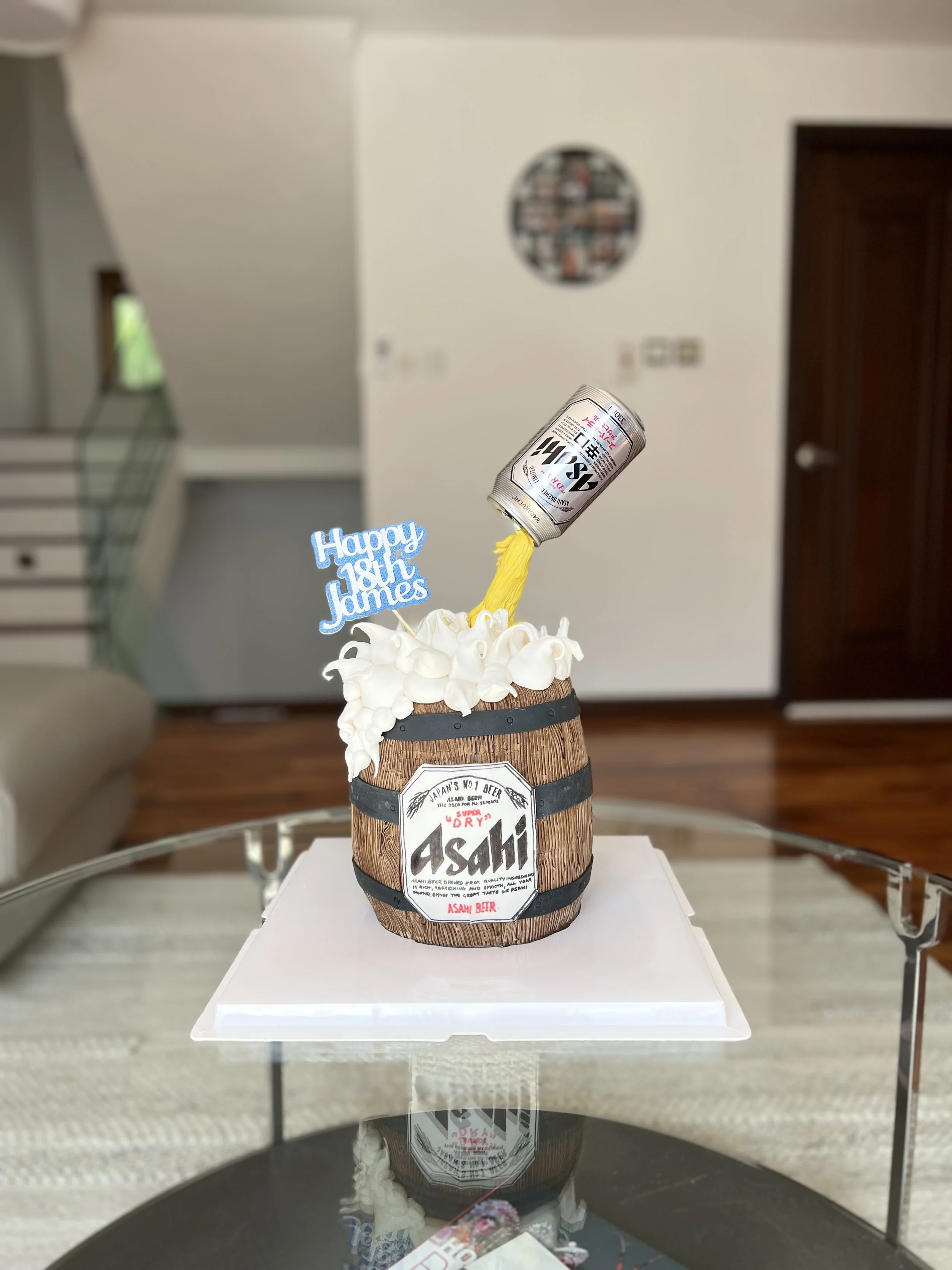 A birthday cake shaped like a beer barrel with whipped cream on top, a sign that reads 'Happy 18th James,' and a can of beer pouring yellow liquid into the barrel, set on a glass table.