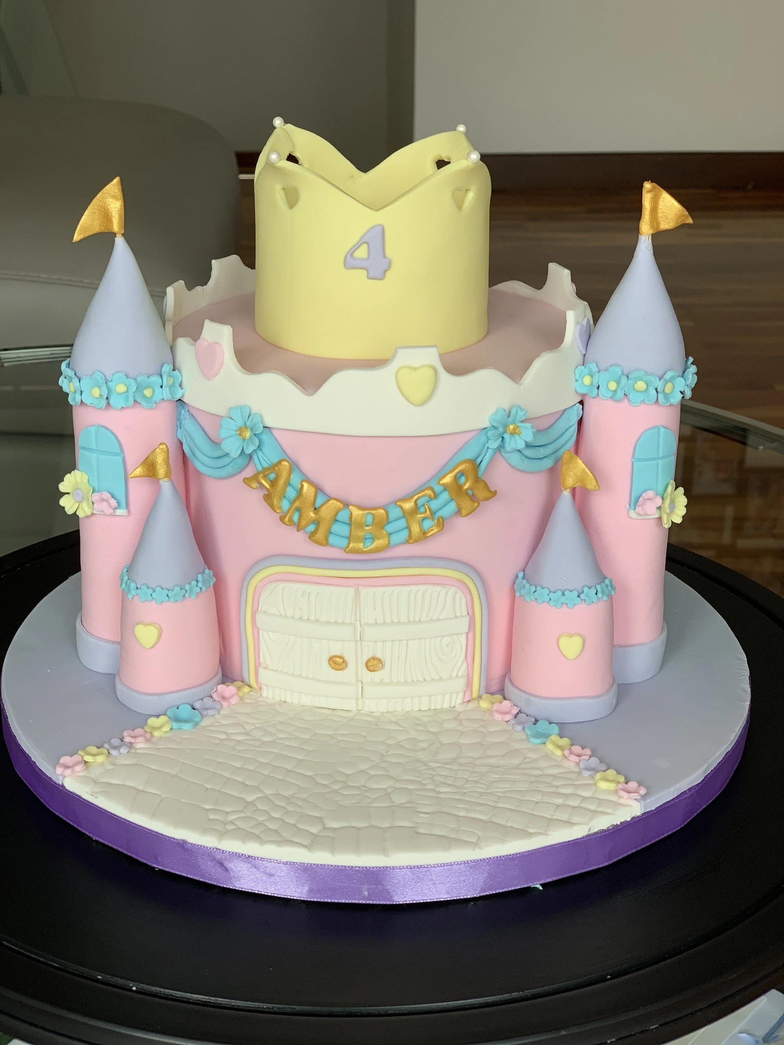 A pink and white castle-themed birthday cake with blue accents and a yellow top. The cake has small castles with flags and a banner spelling 'DAMBER.' A yellow number 4 is on top of the cake.