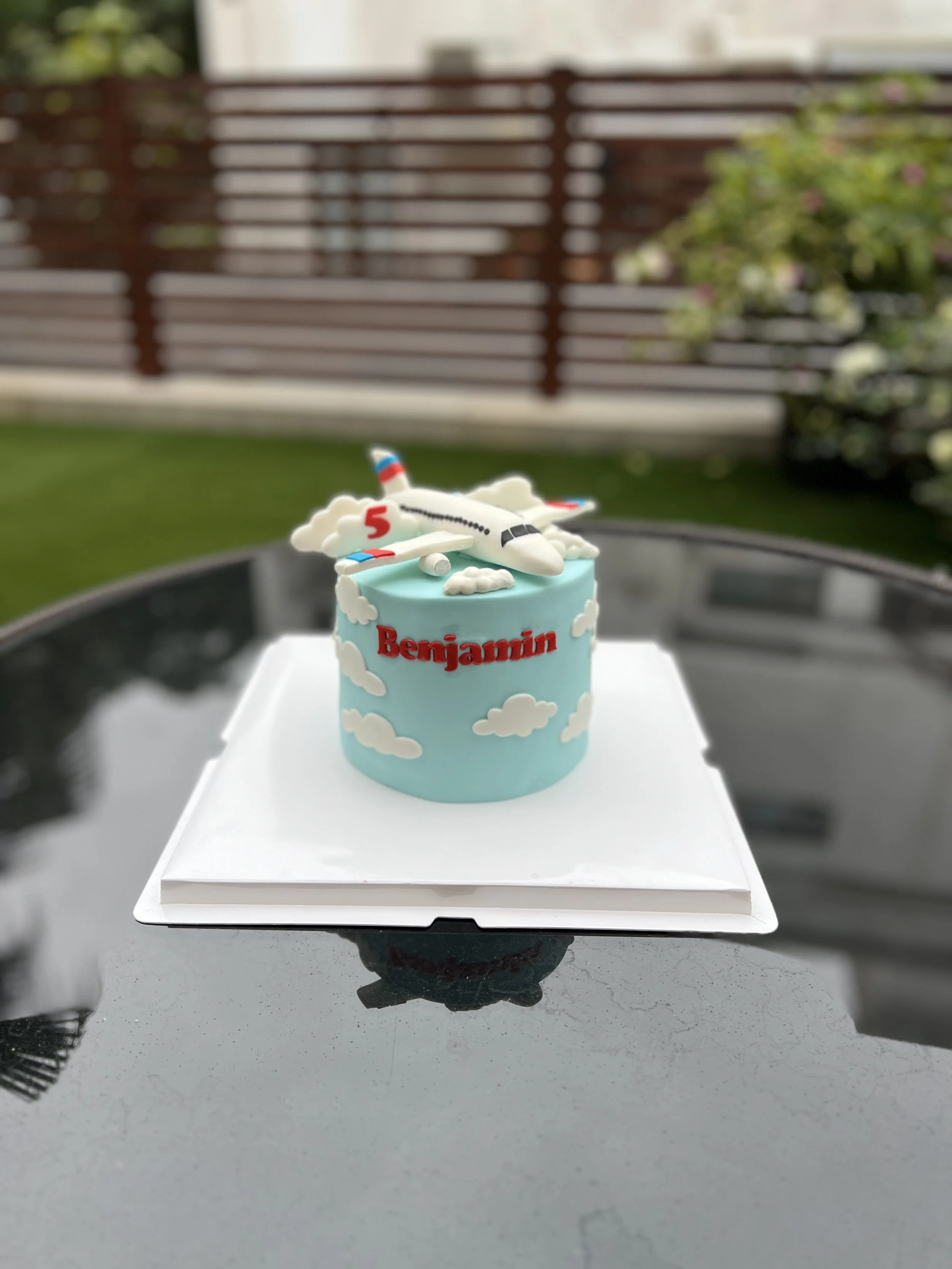 A birthday cake with a white airplane topper and clouds, with the name 'Benjamin' written in red, displayed outdoors on a white box.