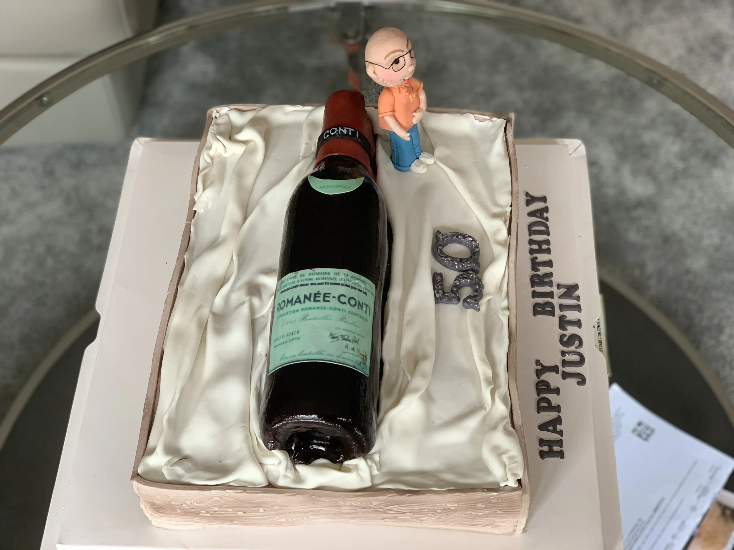 A rectangular birthday cake decorated with white icing, featuring a toy figure of an elderly man, a small figurine of a woman with glasses, a large edible wine bottle, and the words "HAPPY BIRTHDAY JUSTIN" written on the side. The cake has the number