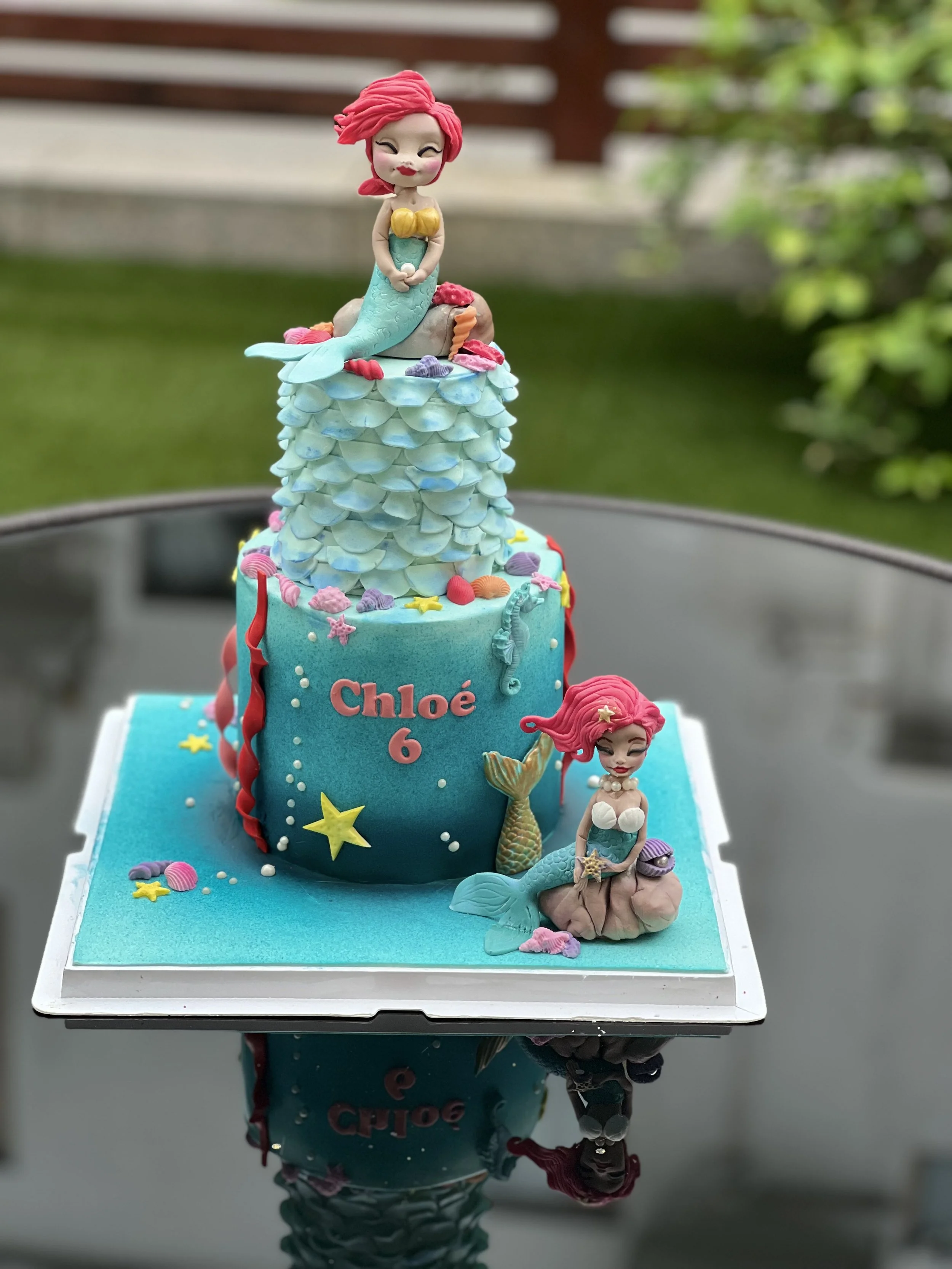 A mermaid-themed birthday cake with two mermaid figurines, colorful seashells, stars, and coral decorations, with the name 'Chloe' and the number 6 on the side.
