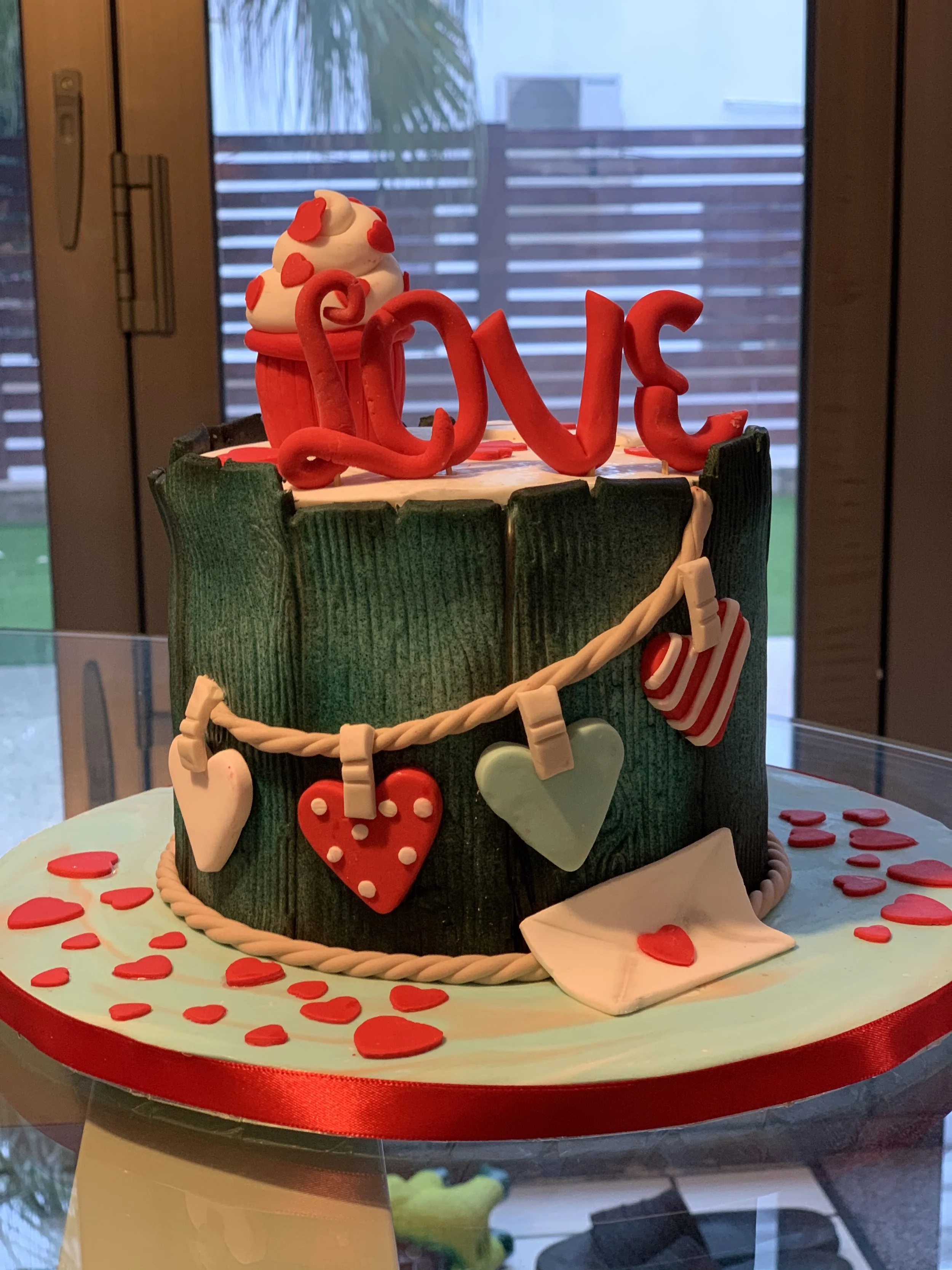 A heart-shaped cake with green textured fondant, topped with red and white decorations. The cake features red letters spelling 'LOVE' on top, a dollop of white frosting with red heart-shaped decorations, and a white fondant envelope with a small red 
