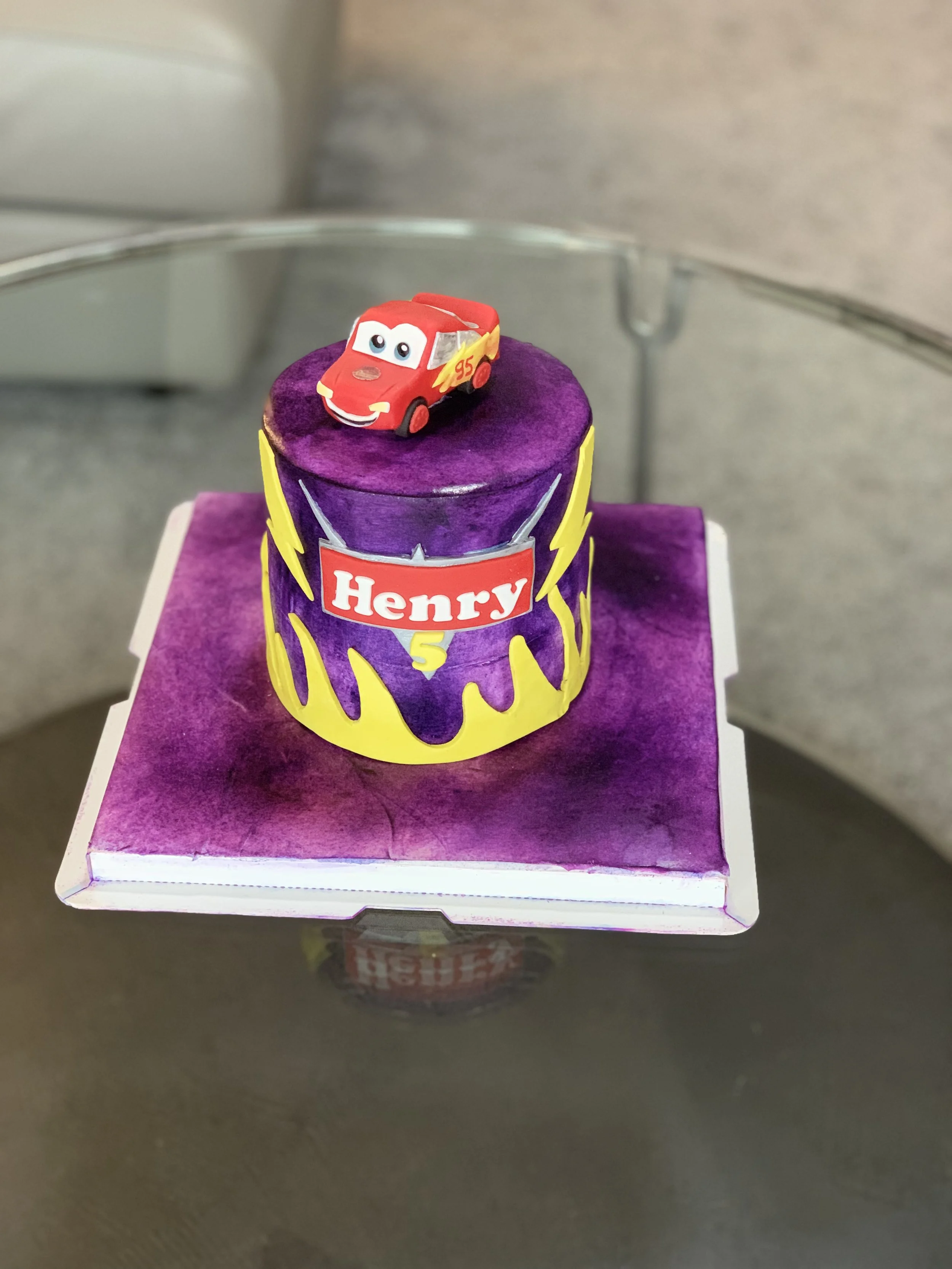 A birthday cake decorated in purple with yellow lightning bolt designs, featuring a Lightning McQueen Toy Story1 car topper on top, with a "Henry" name tag and a yellow number 5.