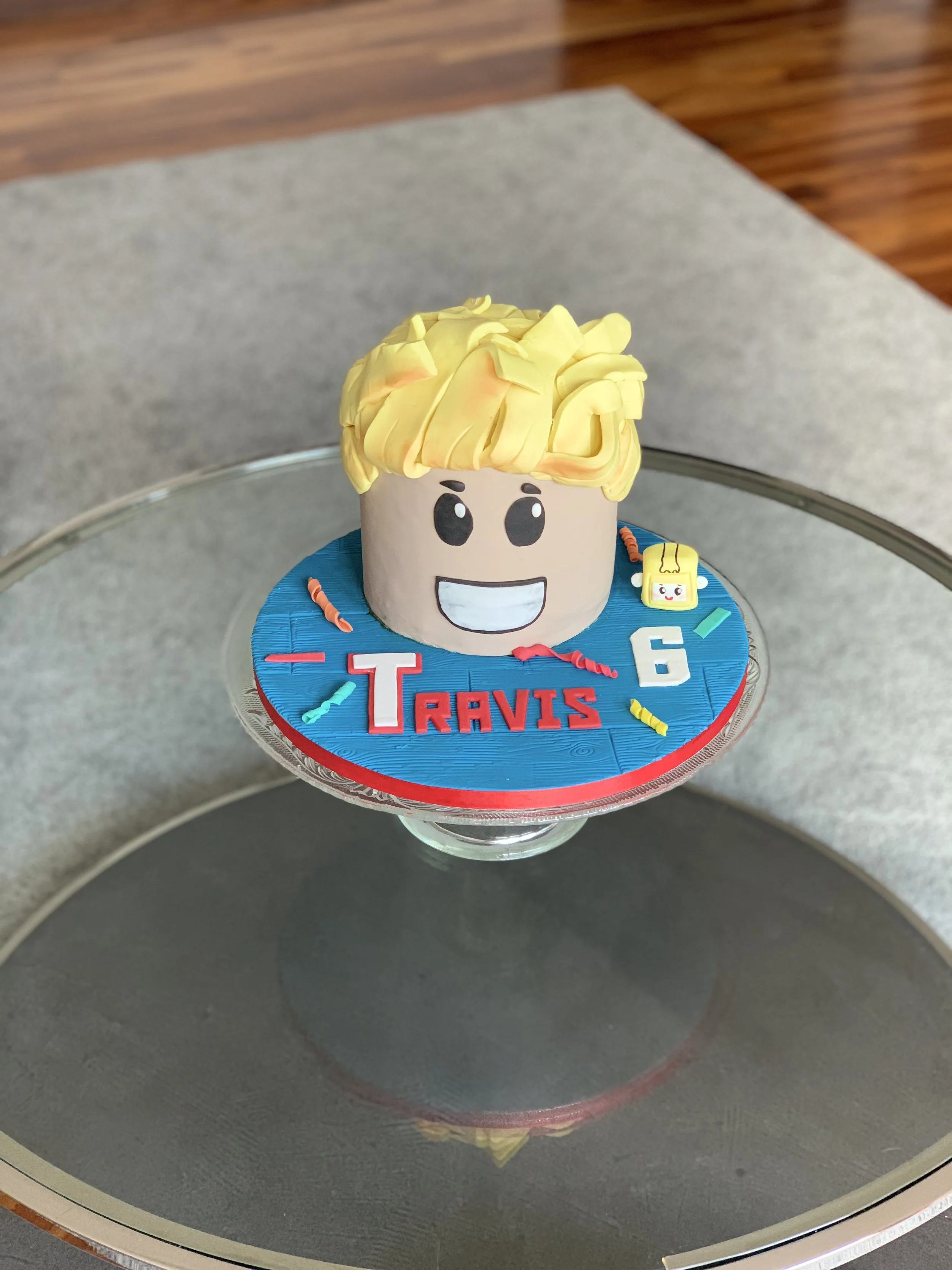 Cake shaped like a cartoon boy's face with blond hair, smiling with big eyes, on a blue and red base with the name 'Travis' and the number '6', decorated with small colorful candies and a small yellow backpack decoration.