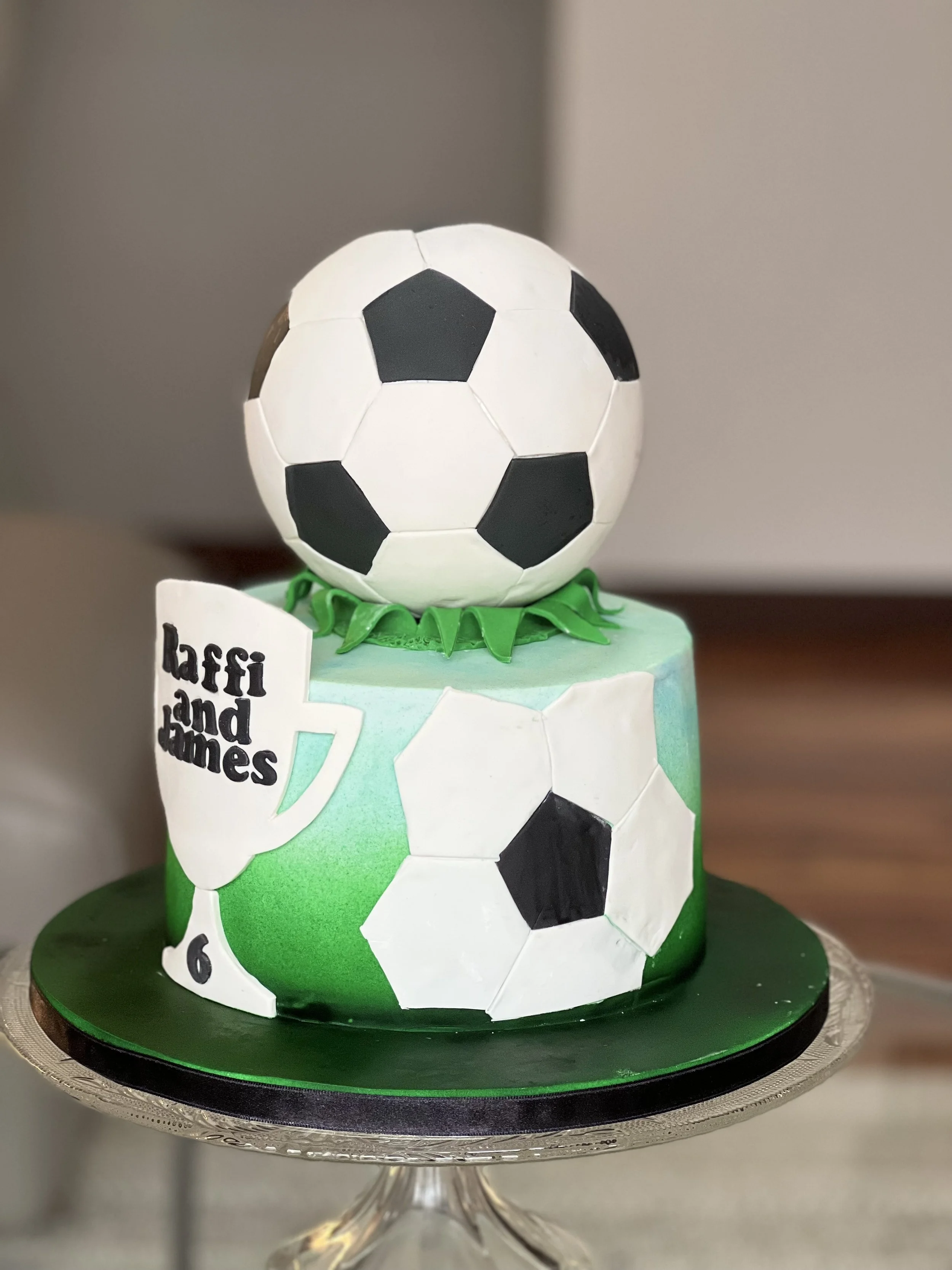 A two-tiered birthday cake decorated with soccer theme. The top tier features a real-looking soccer ball and green grass-like fondant. The bottom tier has soccer ball images, the name "Laffi and James" written on a white cup-shaped decoration, and a 