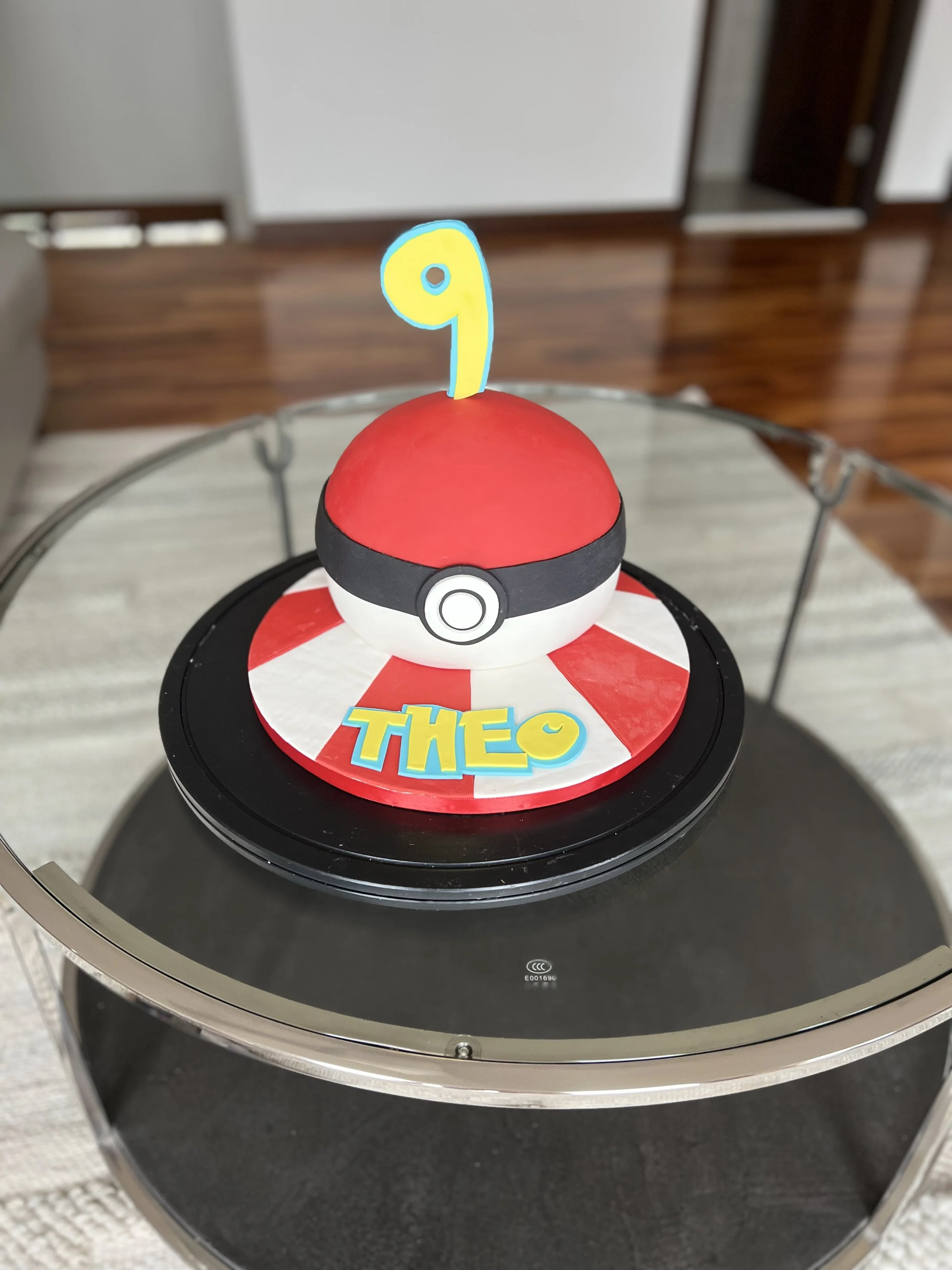 Pokemon-themed birthday cake shaped like a Pokeball with a yellow number 9 candle on top, and a colorful name decoration reading "THEO" in front.