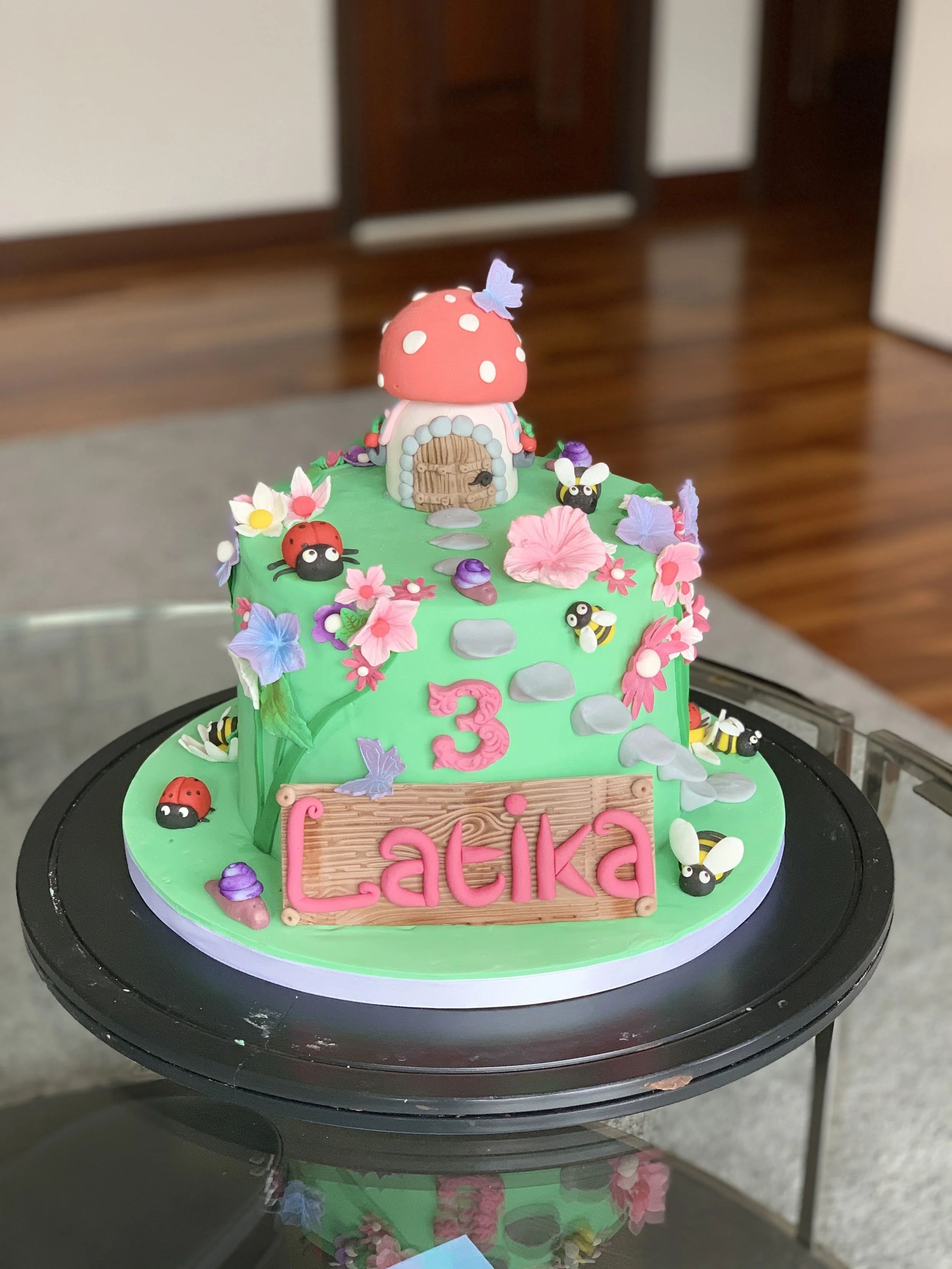 Colorful mushroom-shaped cake decorated with flowers, bees, ladybugs, and butterflies, with a wooden sign reading "Latika" and a pink number three, celebrating a third birthday.