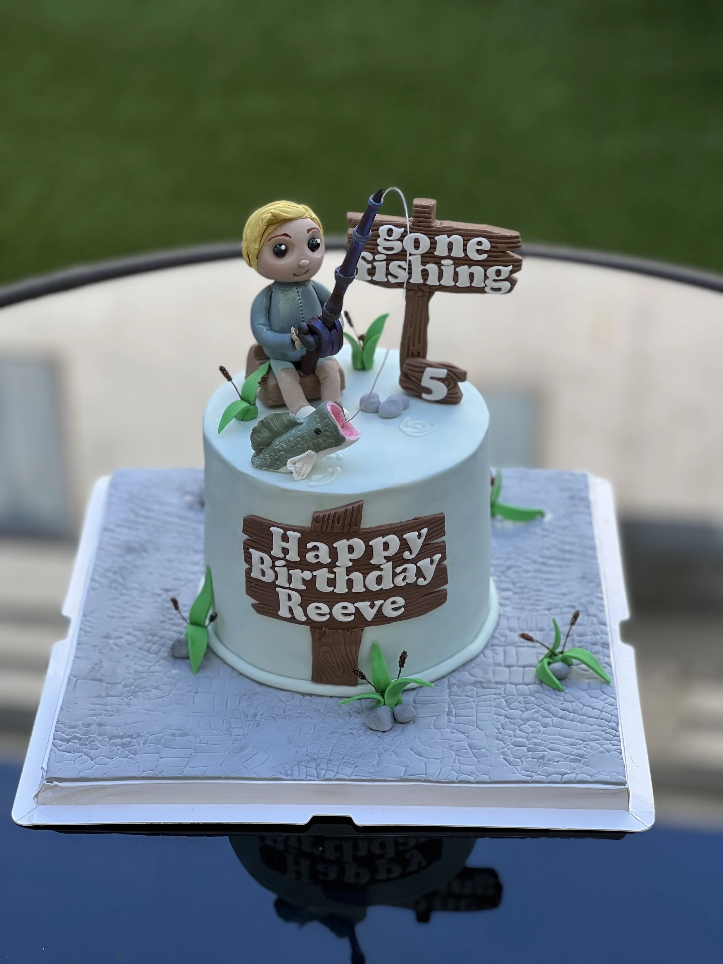 A birthday cake with fishing-themed decorations and a topper of a figurine of a child fishing, surrounded by a fish and green plants. The cake has a sign that says "gone fishing" and another that says "Happy Birthday Reeve."
