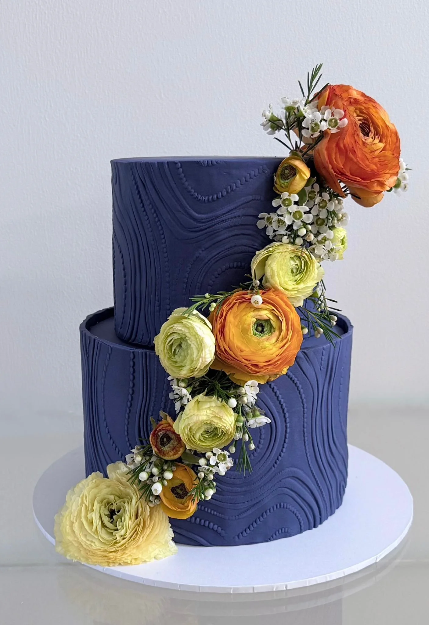 Two-tiered blue cake with textured wavy patterns, decorated with a cascading arrangement of orange, yellow, and cream ranunculus flowers, and small white filler flowers with greenery.