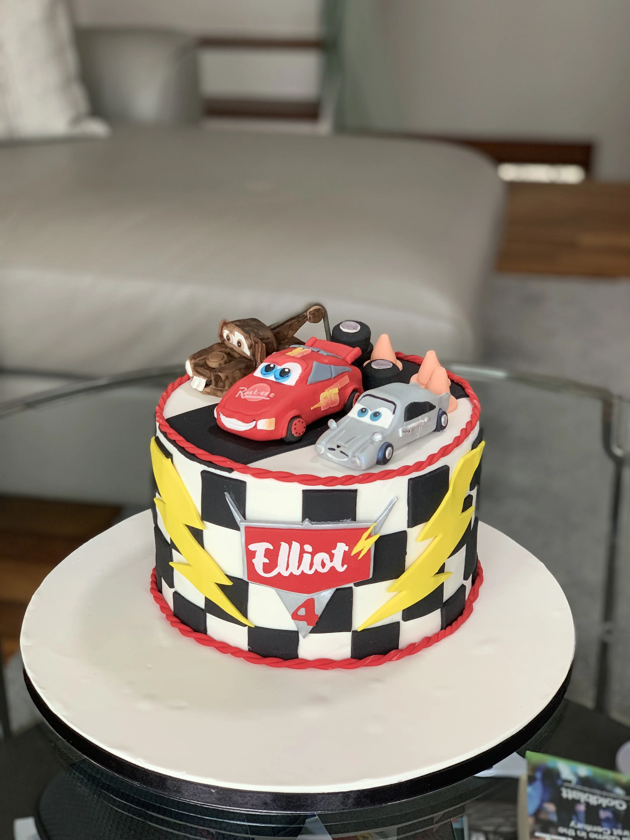 A colorful race car themed birthday cake decorated with Lightning McQueen and Jackson Storm cake toppers, on a white cake stand. The cake has black and white checkered patterns with yellow lightning bolts and a red banner with the name "Elliot" and t