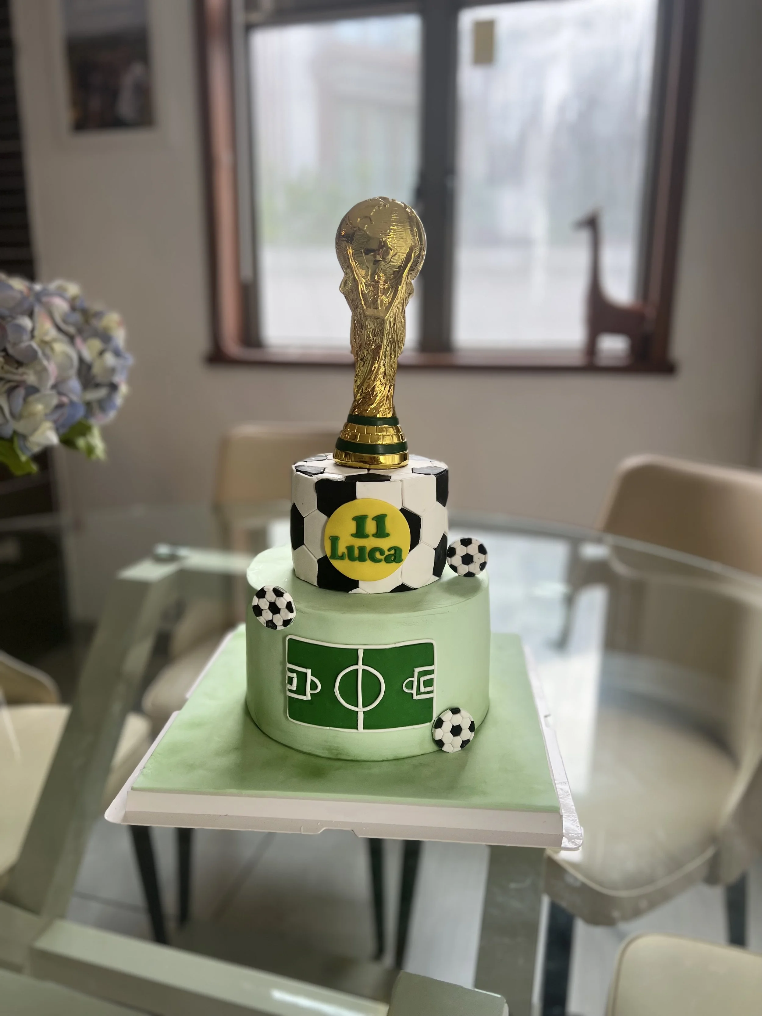 A cake decorated with a football theme, featuring a World Cup trophy topper, soccer ball decorations, and the name 'Luca' on a yellow background, placed on a table in a room with a window and chairs.