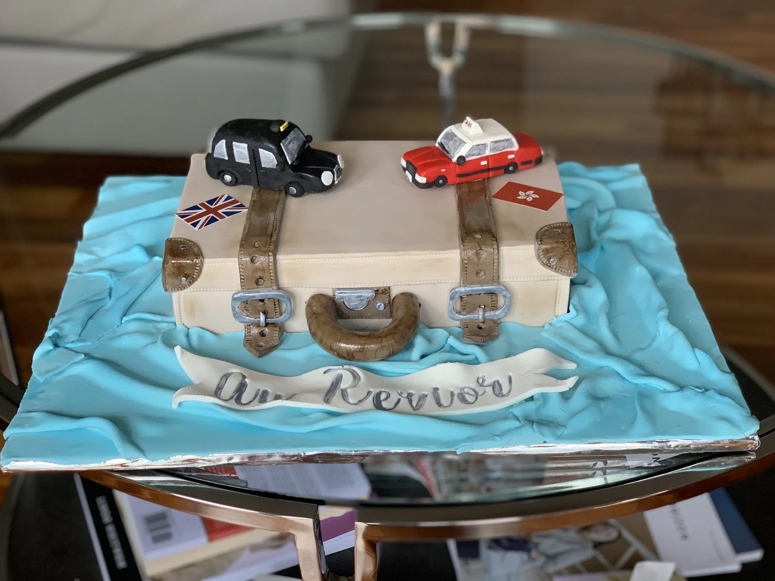 A cake shaped like a suitcase with two toy cars on top, one black and one red, over blue icing that simulates water. The cake has a small British flag and a Hong Kong flag, with a ribbon on the front reading "Air Review."