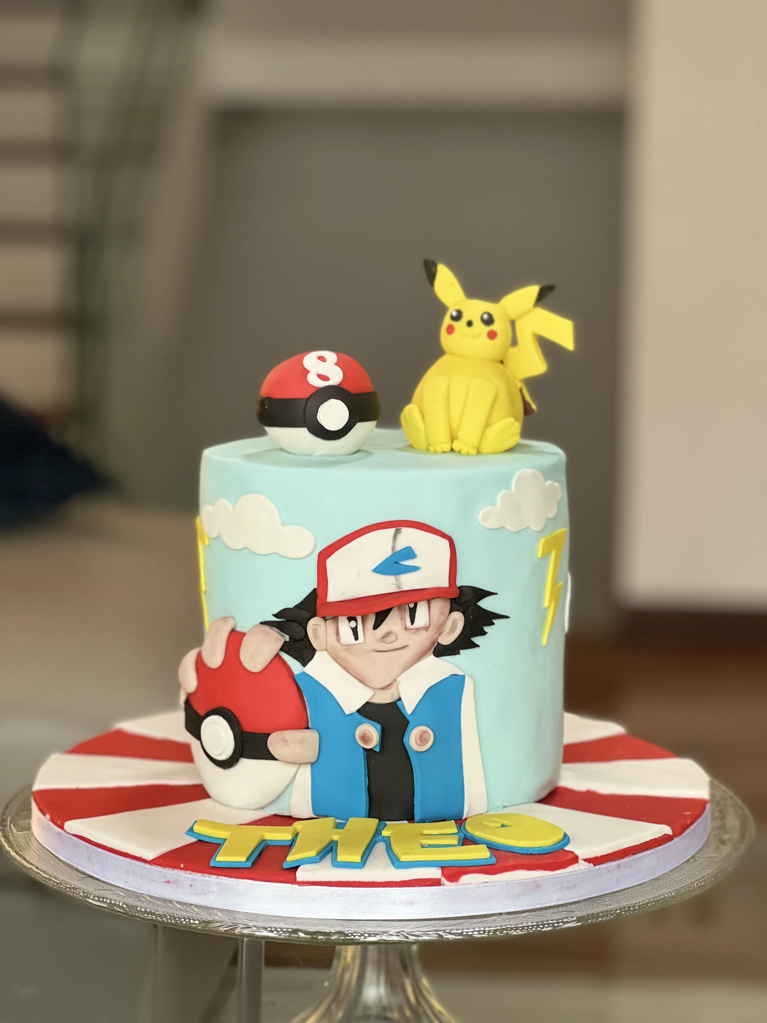 Pokemon-themed birthday cake with Ash Ketchum holding a Pokeball, Pikachu, a Pokeball with the number 8, and the word 'THEO' on the cake base.