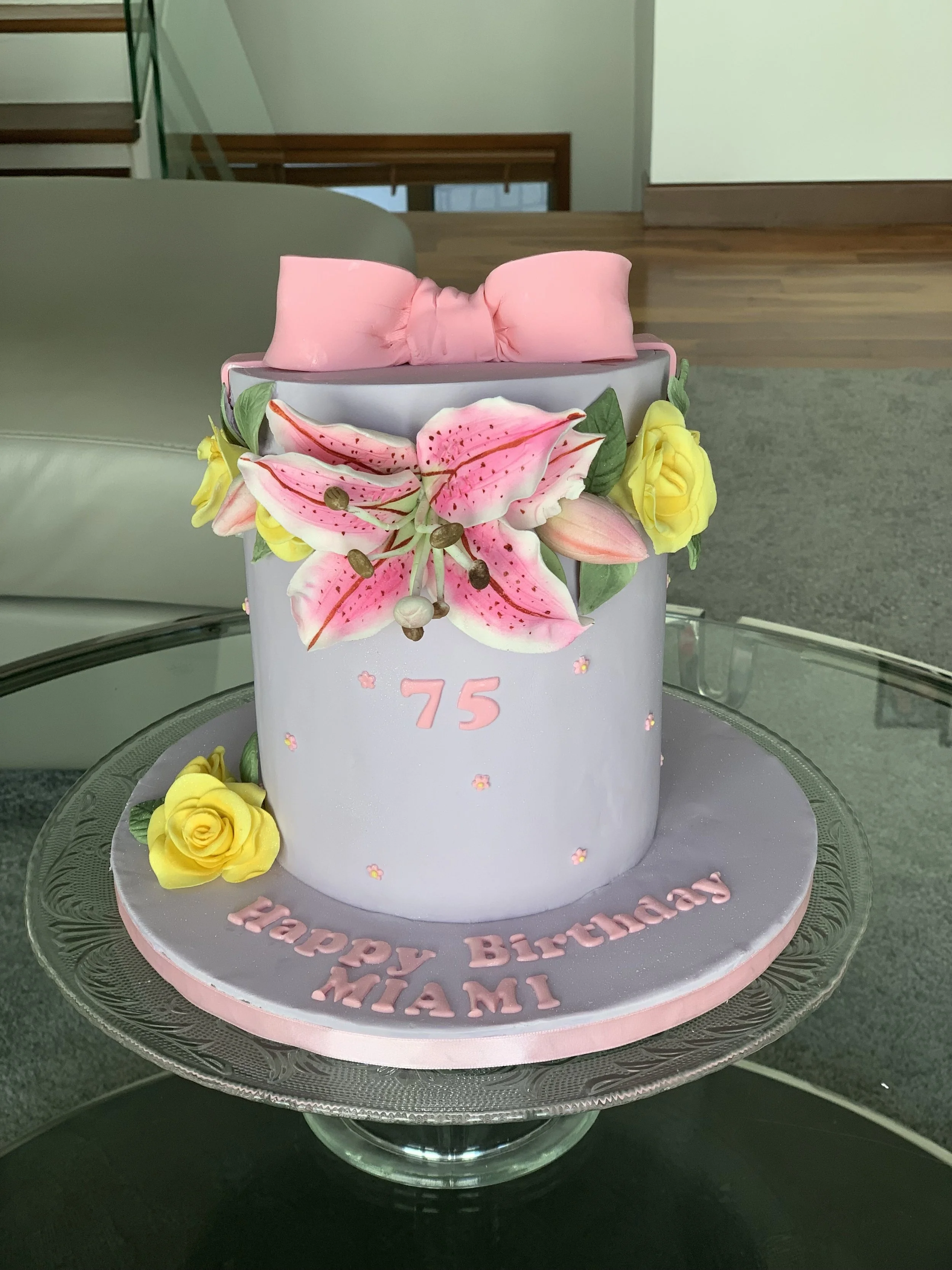 A birthday cake decorated with pink and yellow flowers, a pink bow on top, with the number 75 and text that reads 'Happy Birthday MIAMI' on the base.