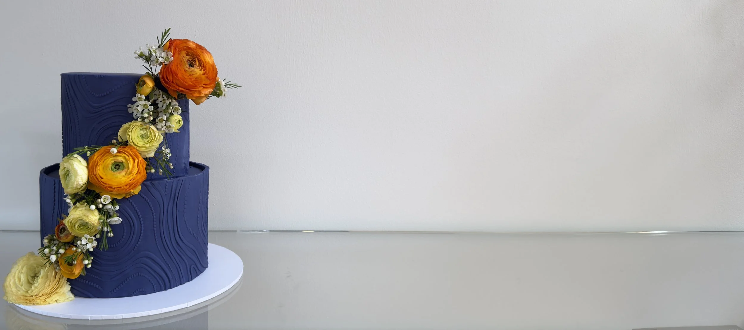 A two-tiered cake with dark blue textured icing decorated with orange, yellow, and white flowers and greenery, placed on a white round cake board on a reflective surface.