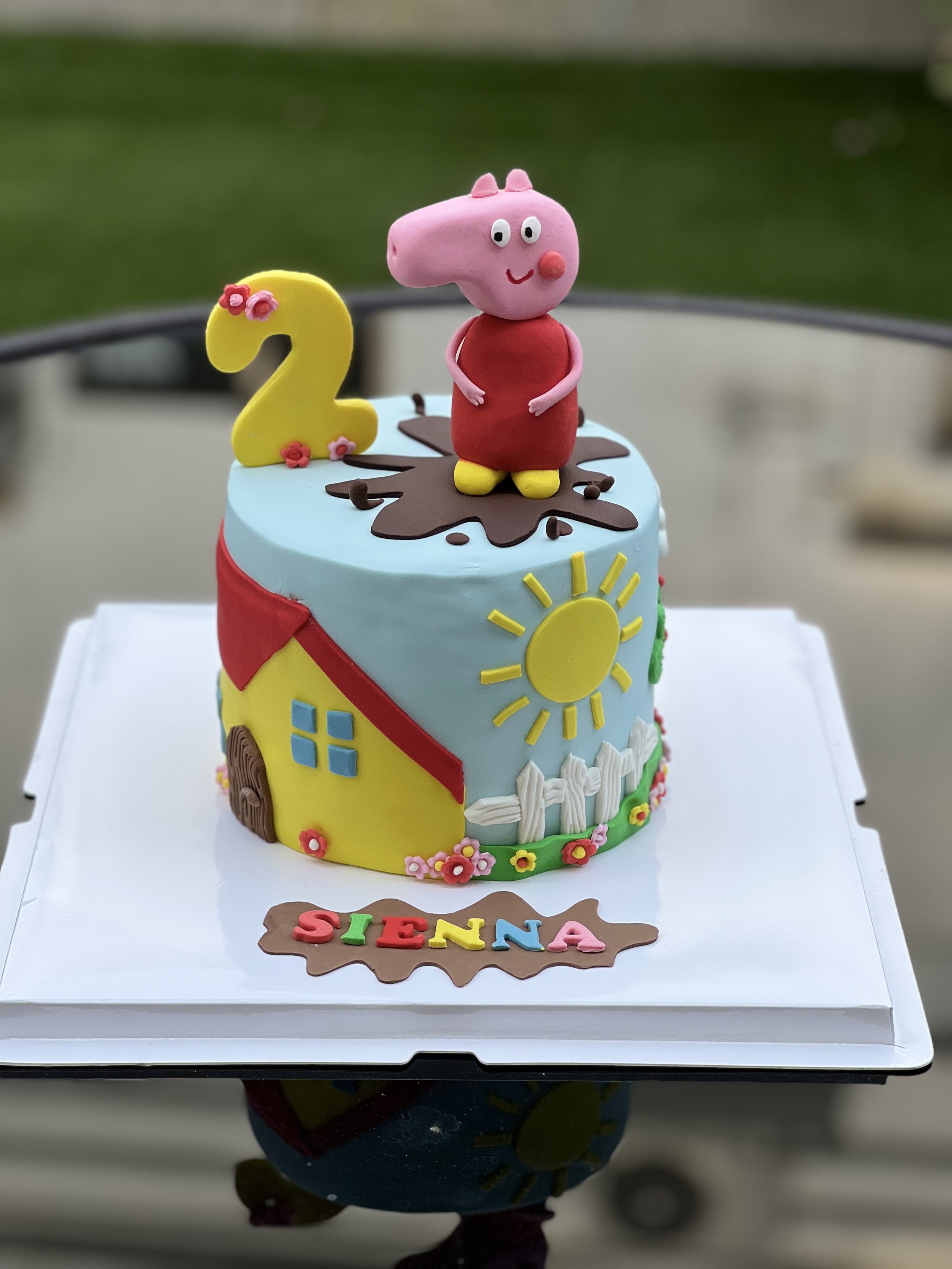 A colorful birthday cake decorated with characters from Peppa Pig, a yellow house, and a sun, celebrating a child's second birthday named Sienna.