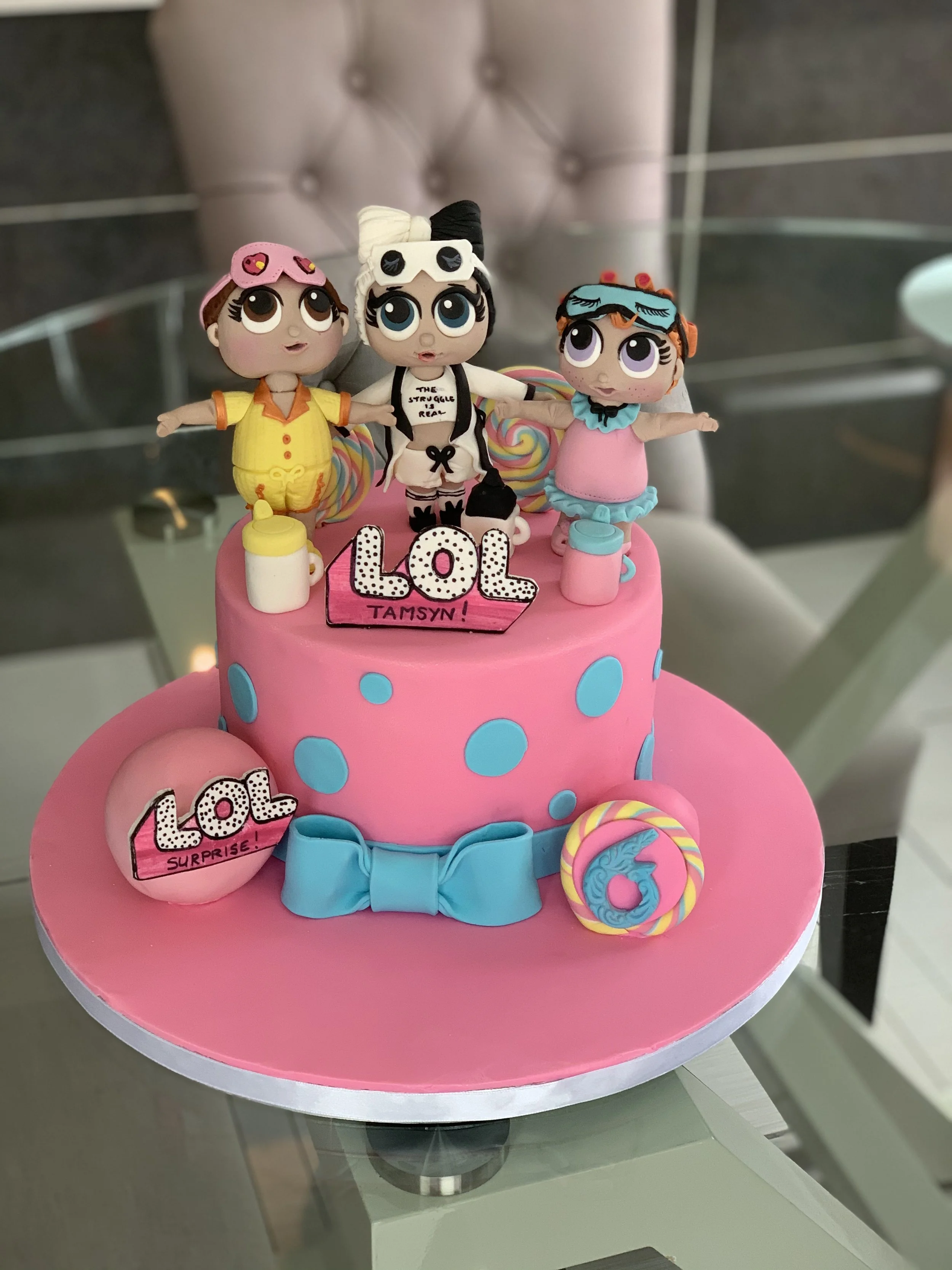 A pink birthday cake decorated with three 'L.O.L.' surprise doll figurines on top, colorful candies, a blue bow, and a pink and yellow swirl lollipop. The cake has blue polka dots and a pink sign reading "L.O.L Tamsyn!"