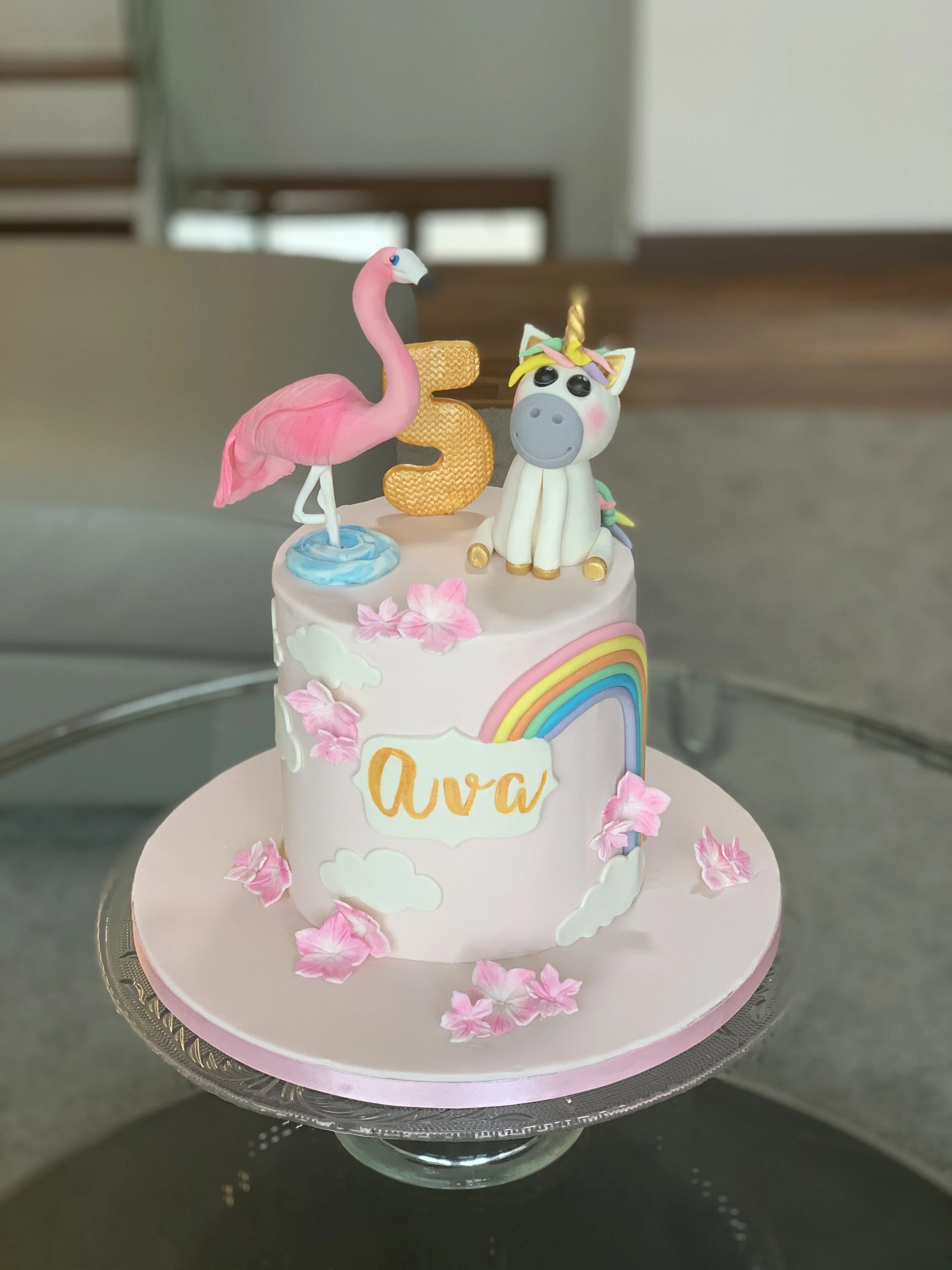 A pink birthday cake decorated with a flamingo, a unicorn, a golden number five, the name 'Ava', a rainbow, clouds, and pink flowers.