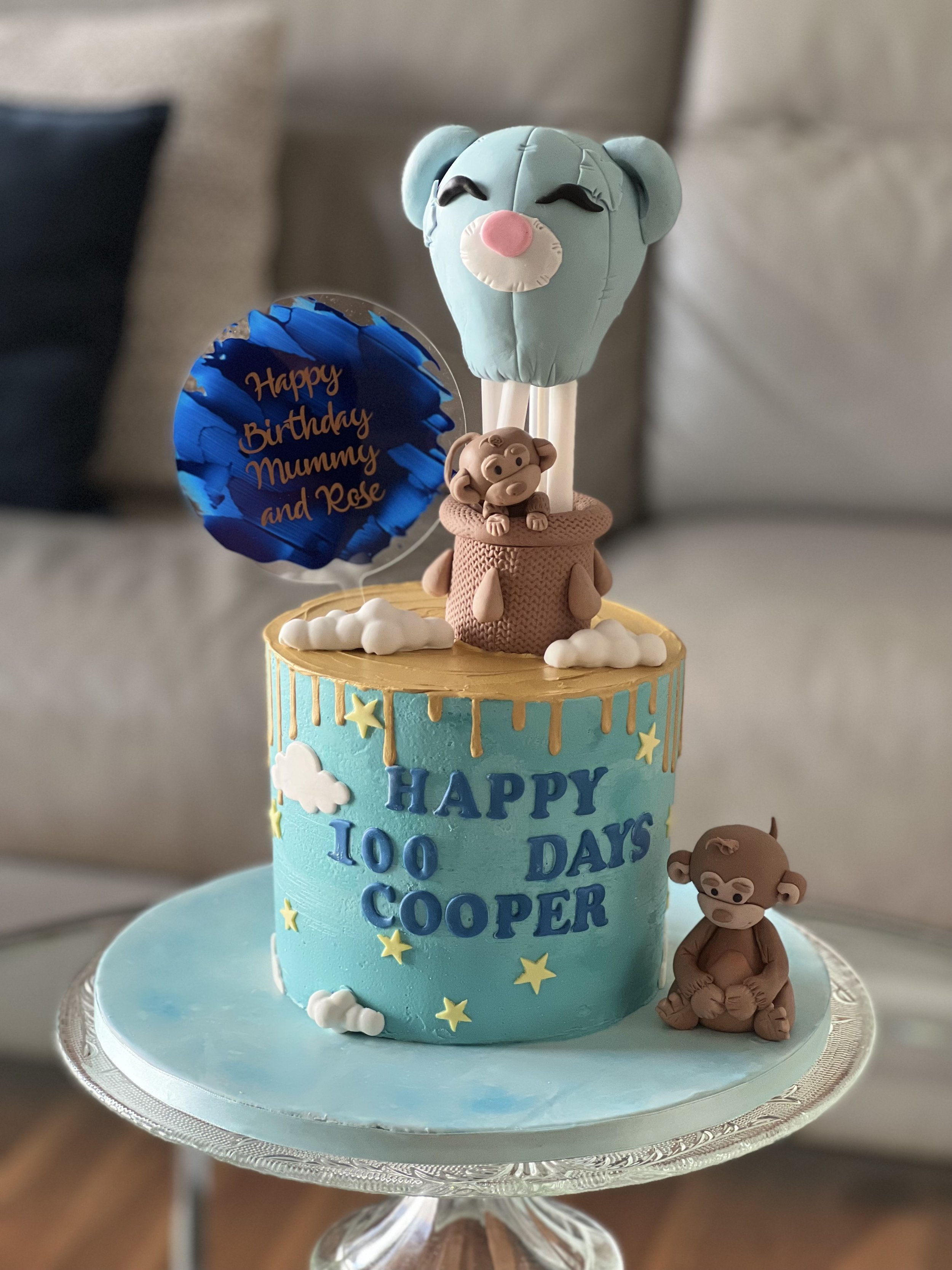 A birthday cake decorated with blue, yellow, and white clouds and stars, celebrating 100 days for Cooper. The cake has fondant animal figures, including a bear on top coming out of a pot, a sitting bear on the side, and a large bear head lollipop top