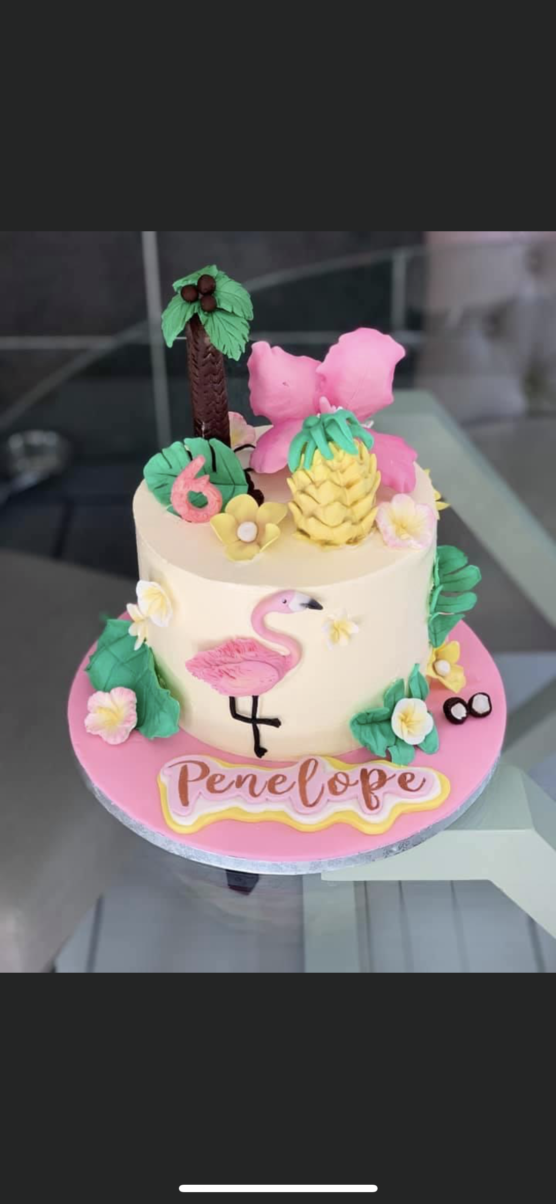 A tropical-themed birthday cake decorated with pink flamingo, pineapple, hibiscus flowers, and banana leaves, with the name 'Penelope' written in pink and yellow on a pink base, and a small '6' indicating the age.