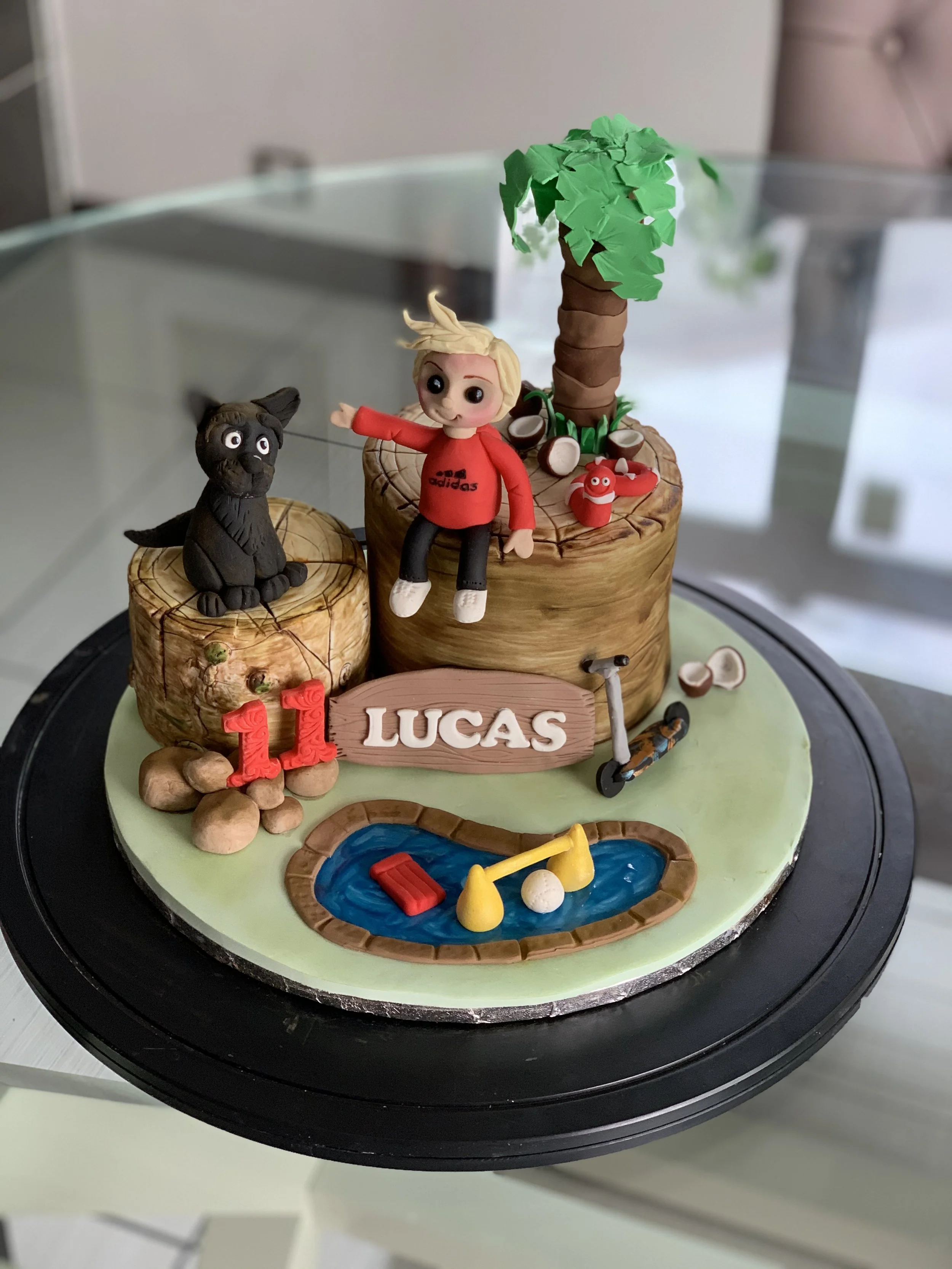A birthday cake decorated with a woody log theme, a small pond, and toy figures, including a boy, a black dog, and a large tree, celebrating Lucas's 11th birthday.