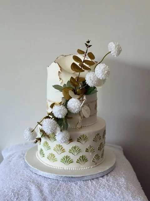 An elegant white two-tier cake decorated with green leaf patterns, topped with a large broken piece of white chocolate or fondant, gold leaves, and white pom-pom flowers, placed on a textured white cloth.