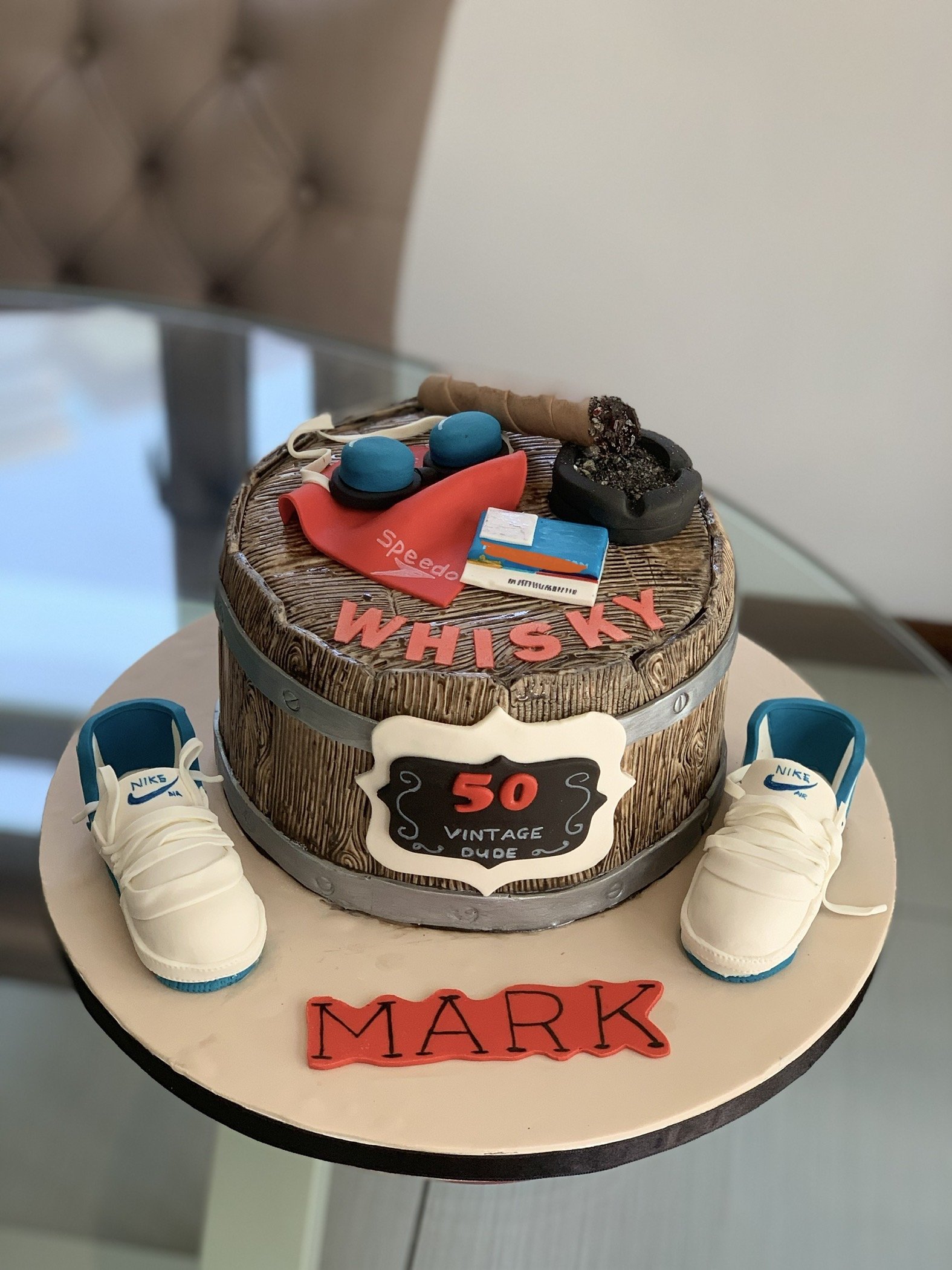 A round birthday cake decorated with a woodgrain pattern, white Nike sneakers, and fondant items including a whisky bottle, cigarettes, and a matchstick. The cake has a sign that reads '50 Vintage Dude' and the name 'Mark' on a red fondant strip.