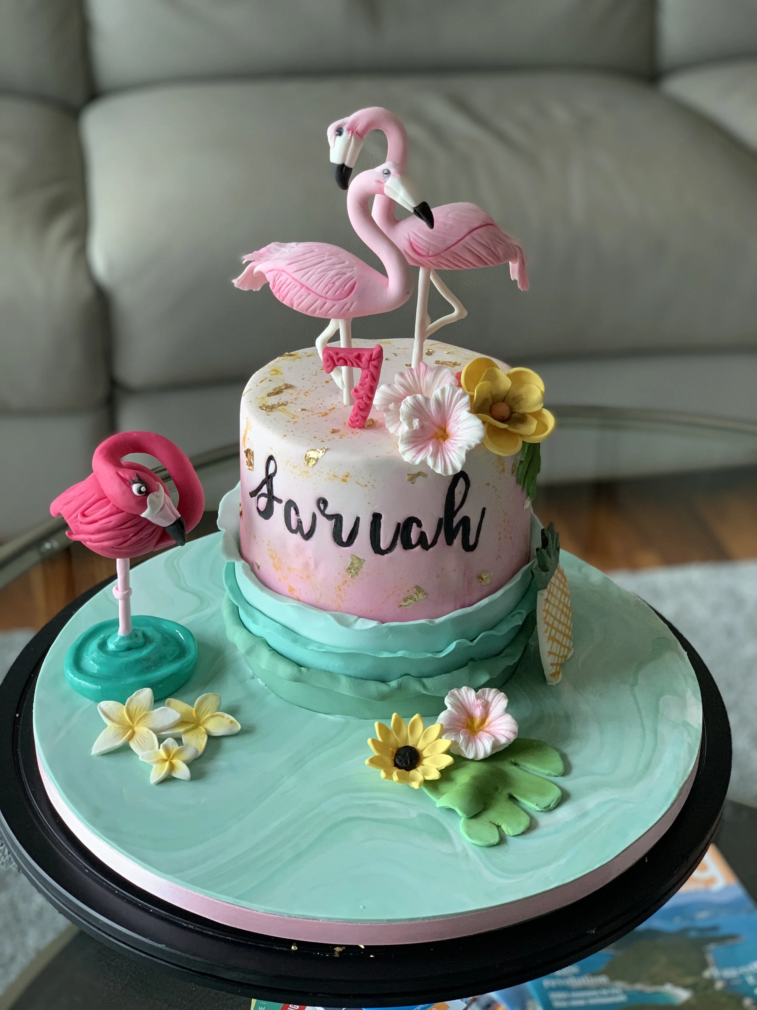 A birthday cake decorated with pink flamingos, flowers, and gold accents, with the name 'Aarav' written on it. The cake has a pink and turquoise color scheme, and the top features two flamingo figures with one holding a number '7' candle.