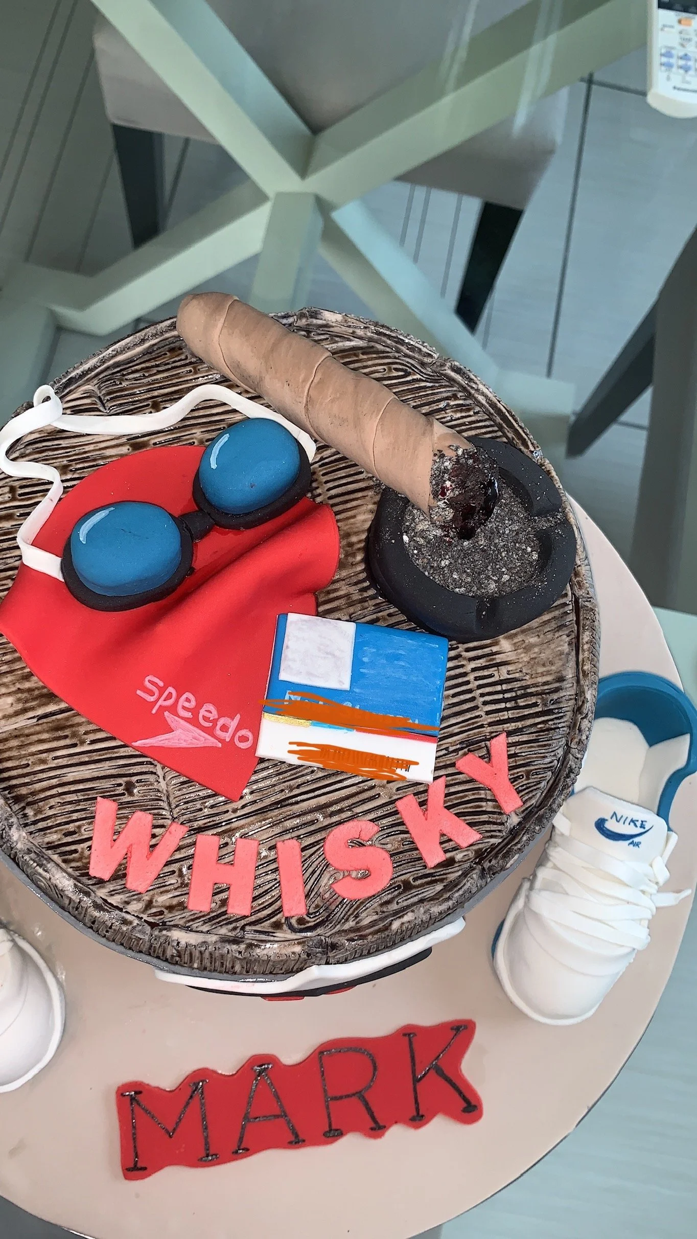 A chocolate cake decorated with a red Speedo cloth, a chocolate cigar, a pair of blue goggles, a small box, and cut-out letters spelling "WHISKEY" and "MARK."