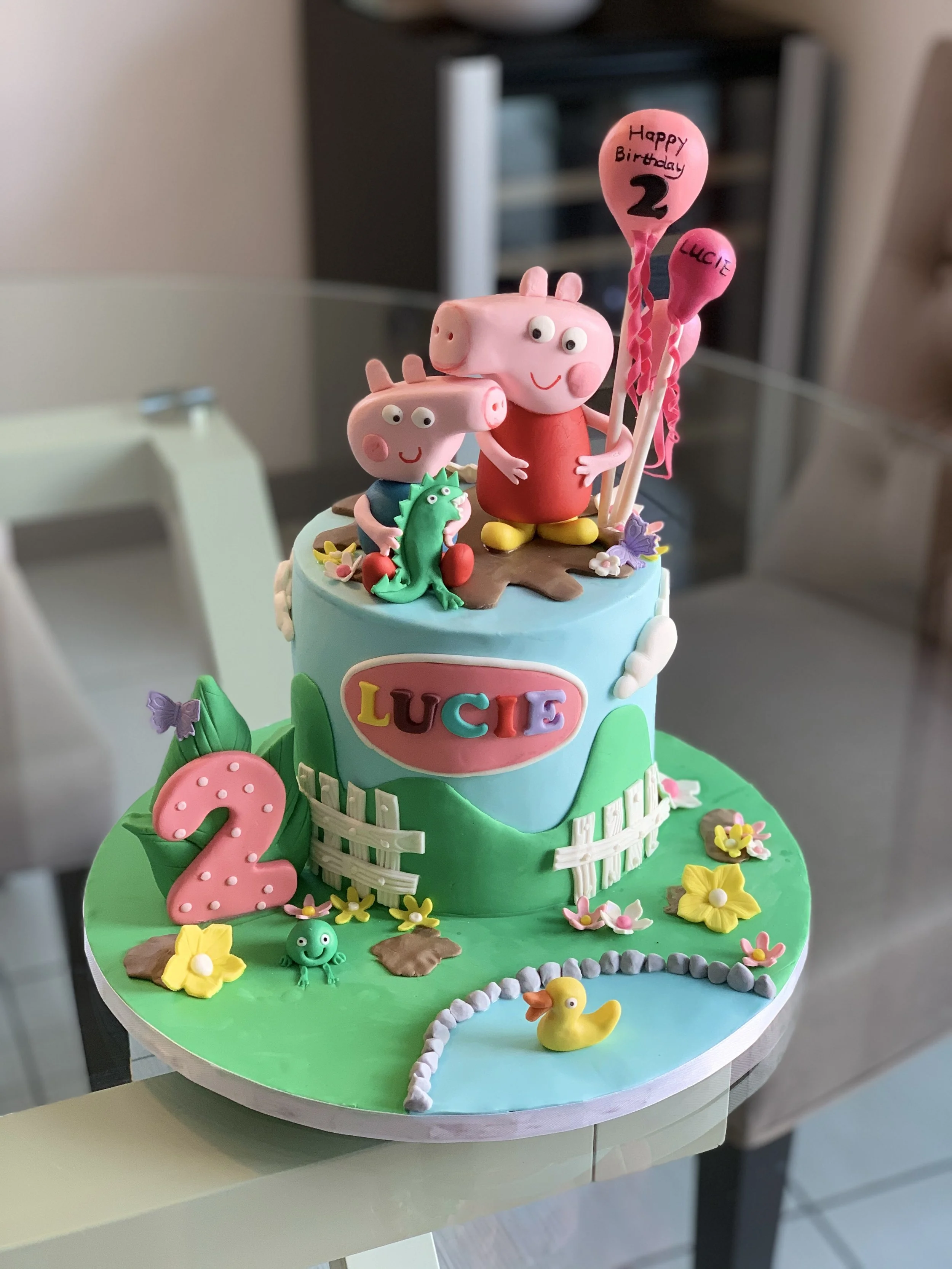 Colorful children's birthday cake with Peppa Pig theme, decorated with Peppa Pig and George figures, a pink number '2' candle, and various flowers, butterflies, and a rubber duck.