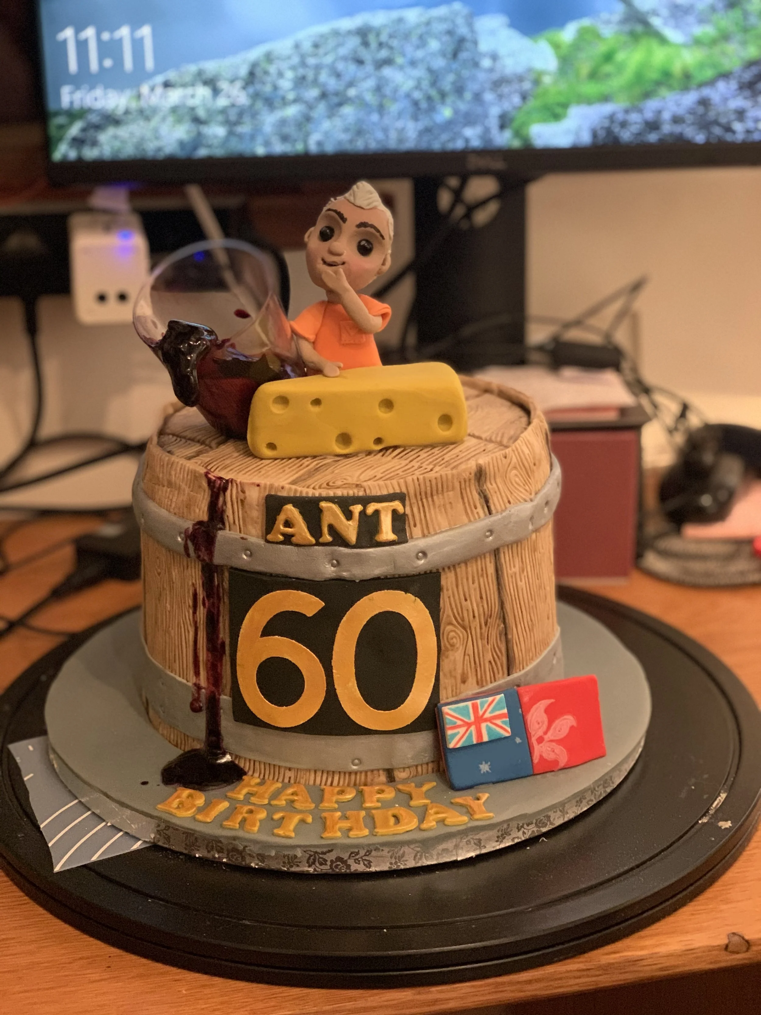 A 60th birthday cake designed to look like a wooden barrel, with a figure of a boy and a glass of wine on top. The cake has the text "ANT 60" and a "Happy Birthday" message at the base, with small flags and a cheese block decoration.