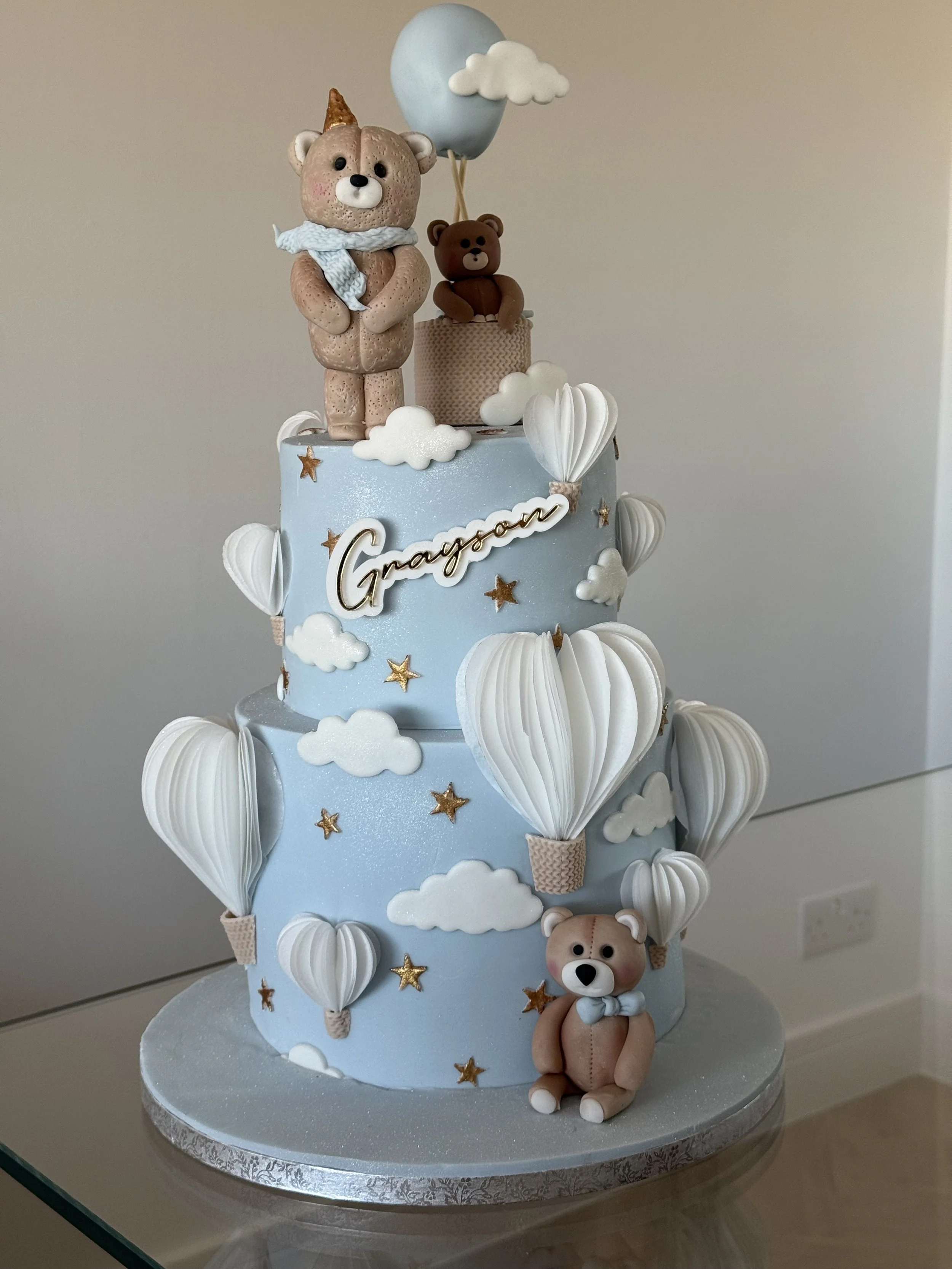 A blue, three-tiered cake decorated with clouds, gold stars, and white hot air balloons. The cake has fondant bears, one on top with a party hat and scarf, and one sitting at the base. The middle tier has a personalized name, "Graydon."