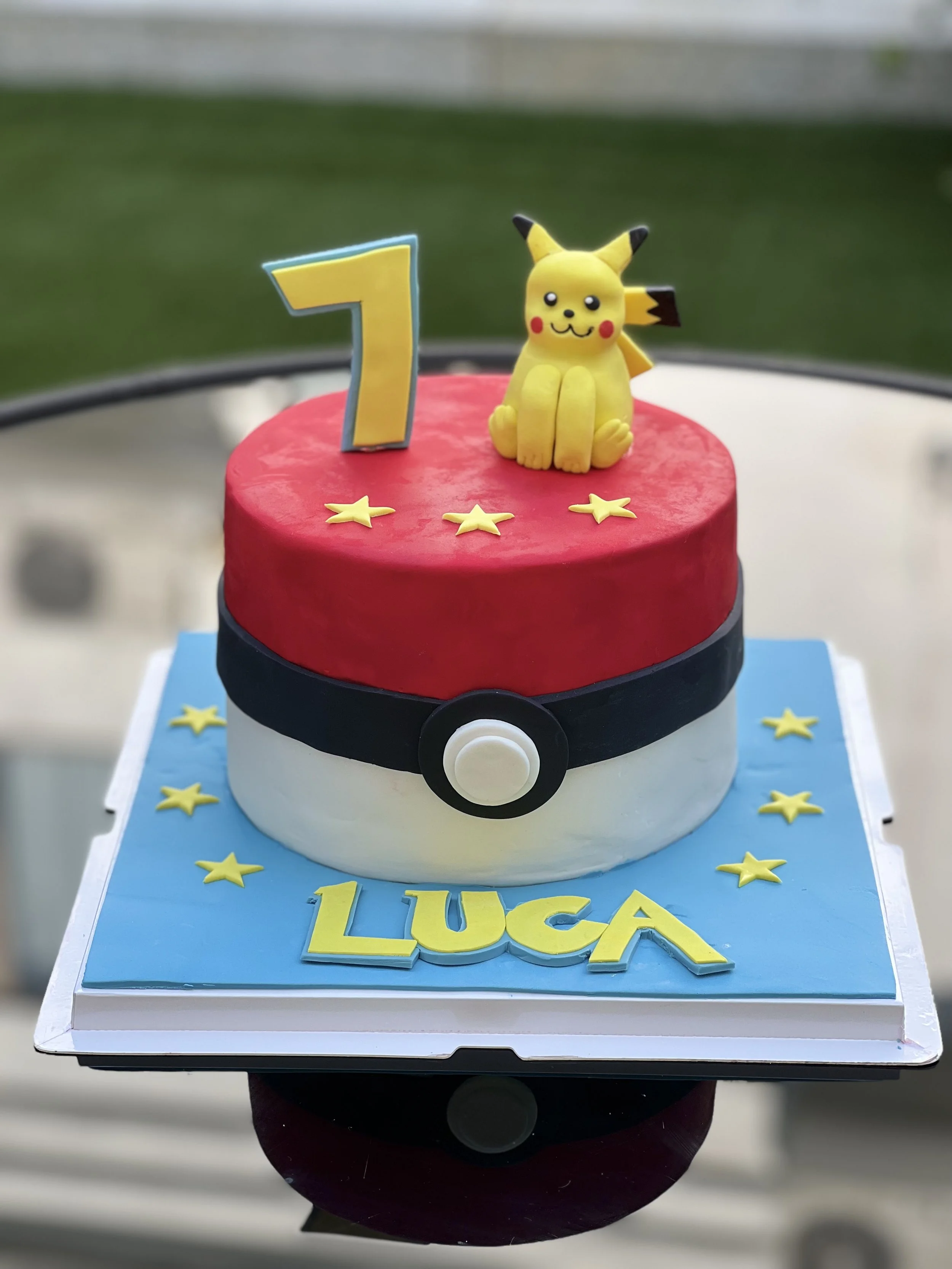 A birthday cake themed after Pokémon with a red top, white bottom, and a black Pokémon Poké Ball design around the middle. The cake is decorated with yellow stars, a Pikachu figurine, and a large number 7 candle. The cake has the name LUCA written on