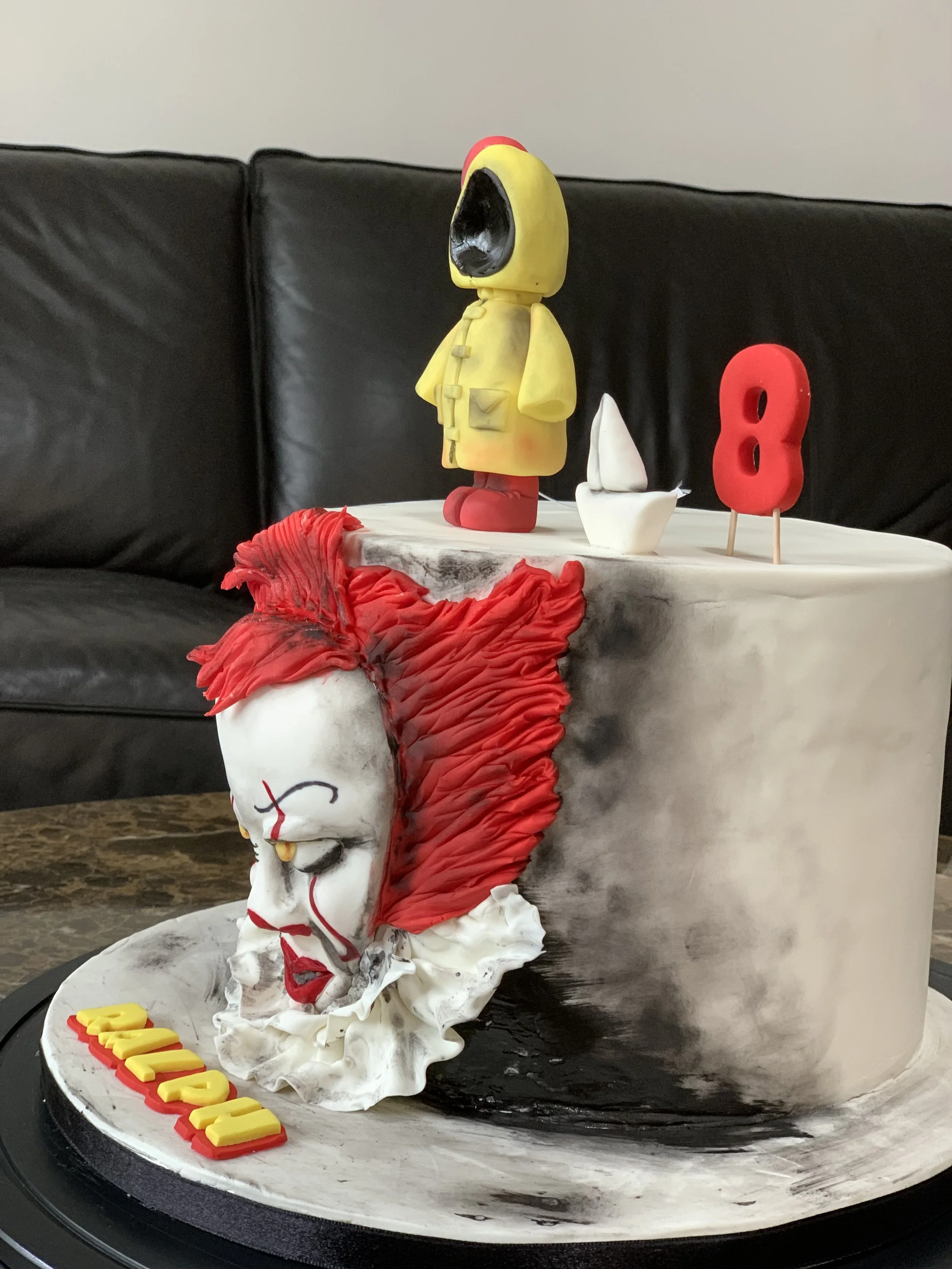 A cake featuring Pennywise the clown from the movie IT, with red hair, a white face, and yellow eyes, next to a small yellow figure wearing a raincoat and red boots, with a small sailboat and a red number 8