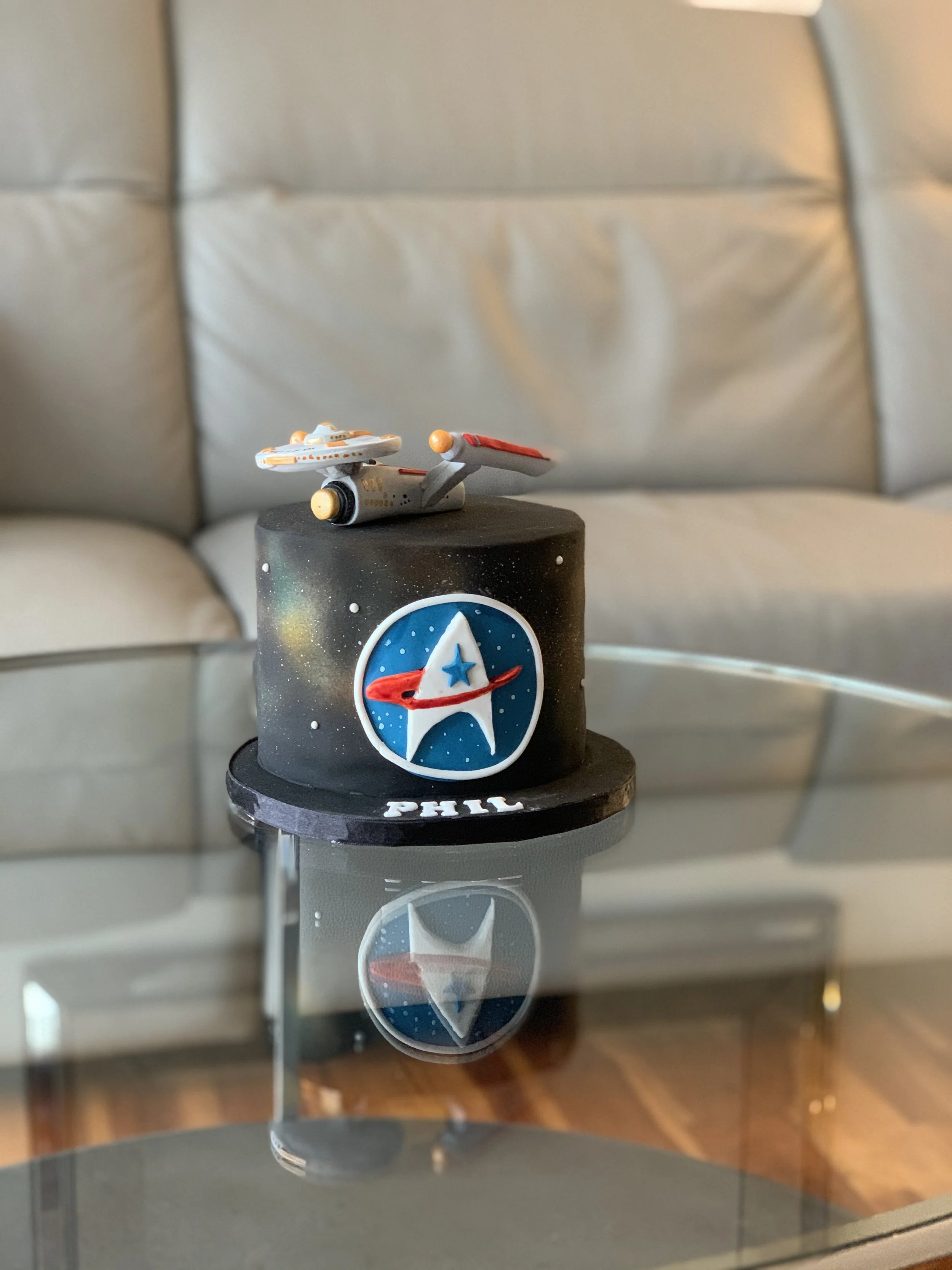 Star Trek-themed cake with a spaceship topper and a 'Star Trek' insignia on the front, placed on a reflective glass table.