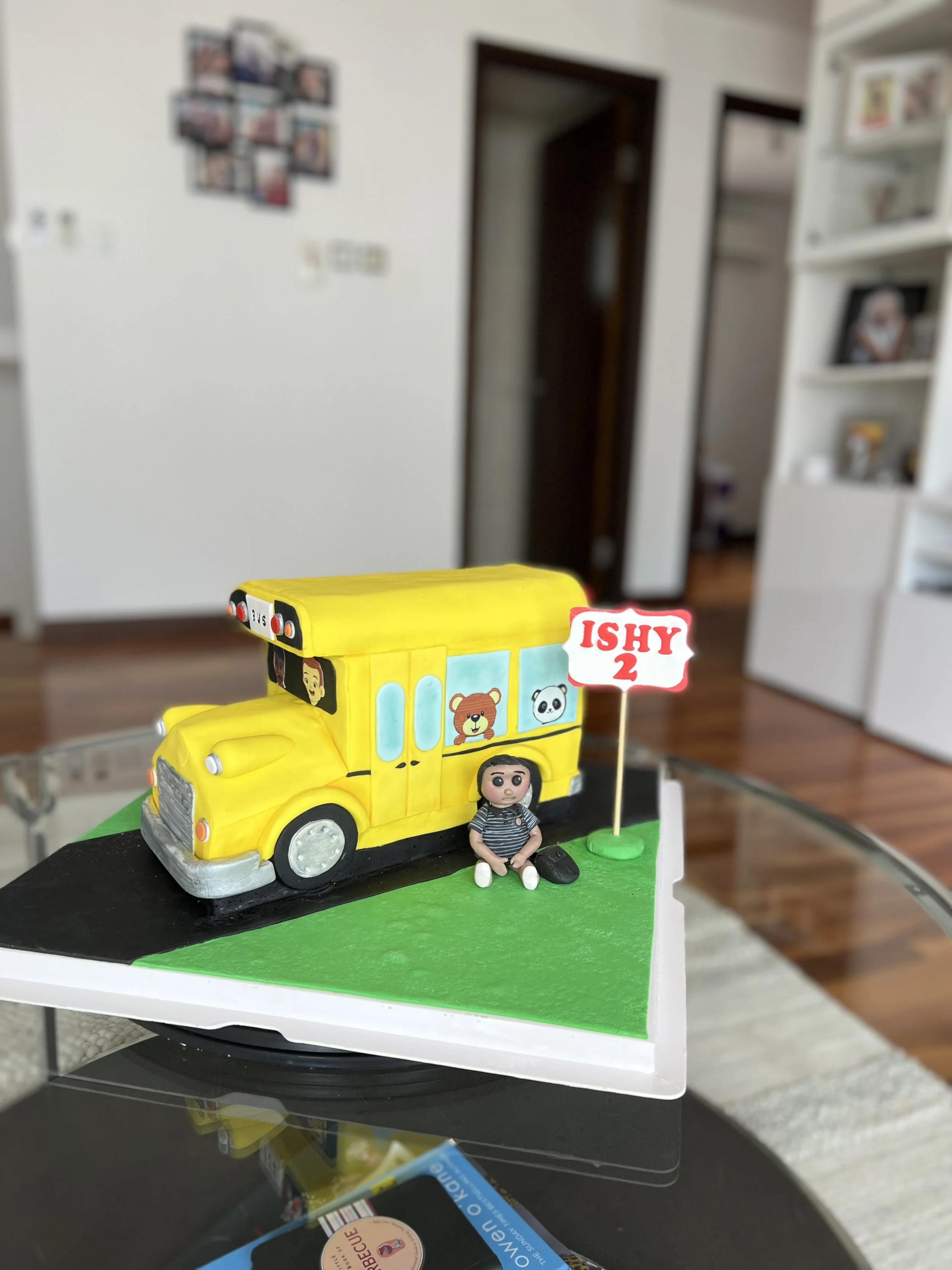 A birthday cake shaped like a yellow school bus with a small figurine of a girl sitting beside it and a sign that reads 'ISHY 2', on a black and white base in a home setting.