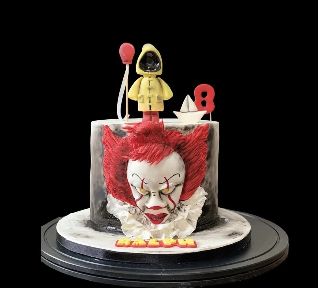 A carnival-themed birthday cake features Pennywise the clown from Stephen King's "It" with red hair and a white face, a small boat, and a yellow raincoat with a red balloon figure on top. The cake has black and white marbled appearance with the word 