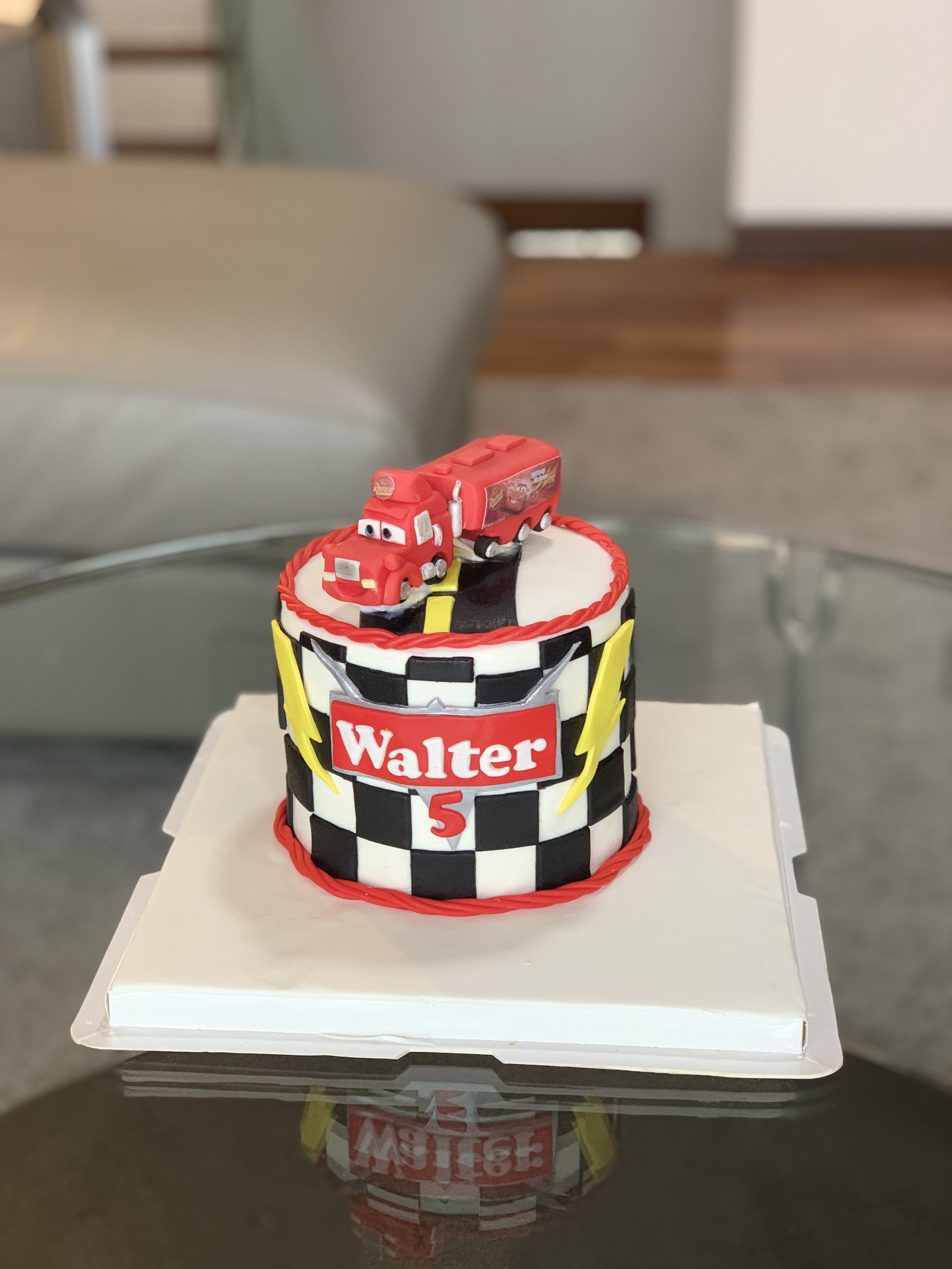 A birthday cake decorated with a racing car theme, featuring a toy race car on top, with the name 'Walter' and the number '5' on the front, and black and white checkered pattern on the sides.