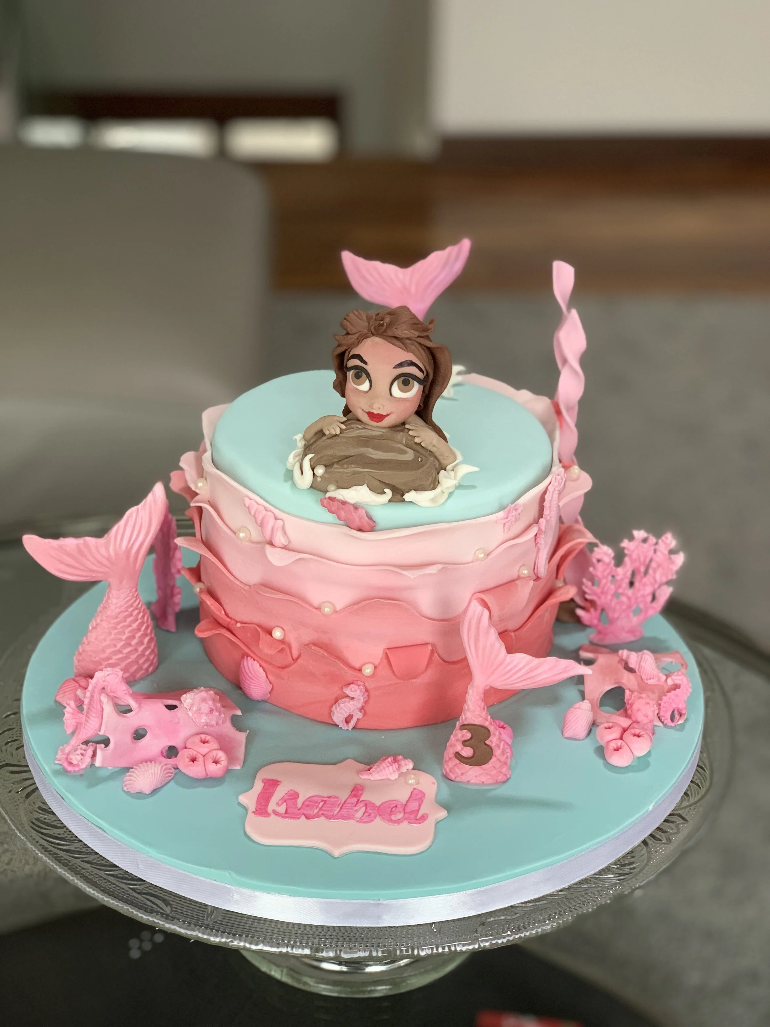 A pink and light blue birthday cake with a mermaid theme, decorated with mermaid tail, seashells, and coral, featuring a mermaid figurine on top, and a pink plaque with the name 'Isabel' and number '3'.