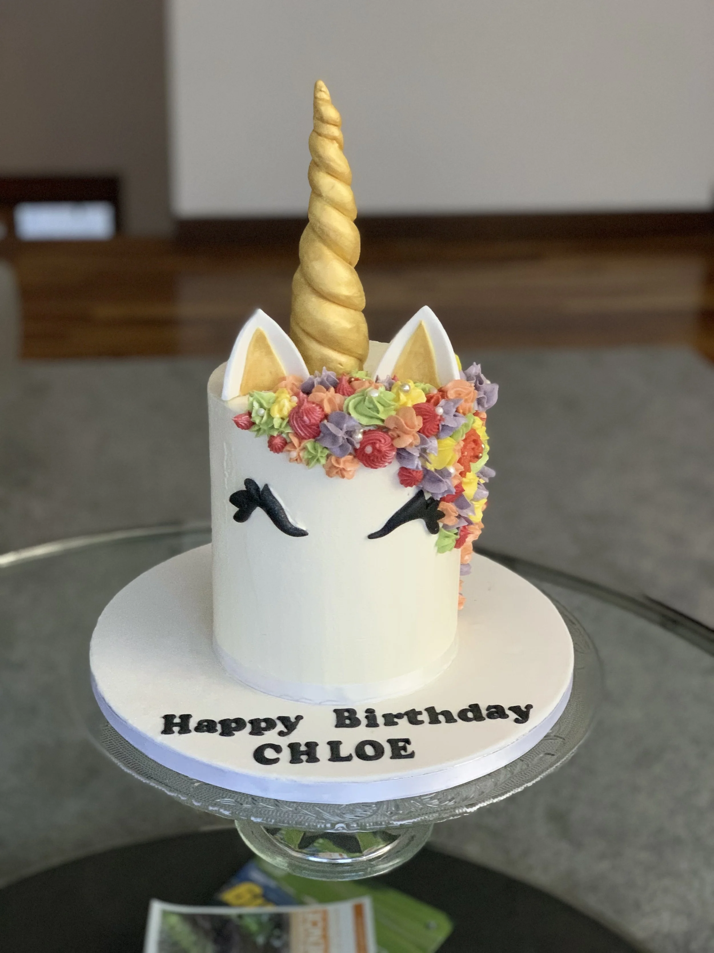 Unicorn-themed birthday cake with a gold horn, pink and purple flowers, and closed eyes, with the message "Happy Birthday CHLOE" on the base.