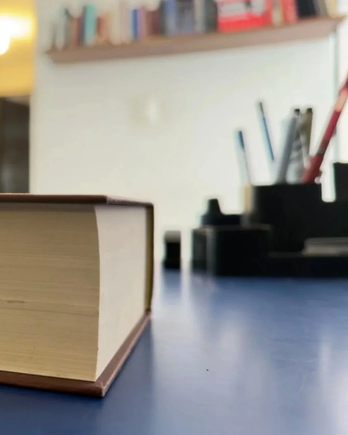 Un libro sobre una mesa con una pila de libros y un organizador de escritorio con varios bolígrafos y el fondo de una estantería con libros.