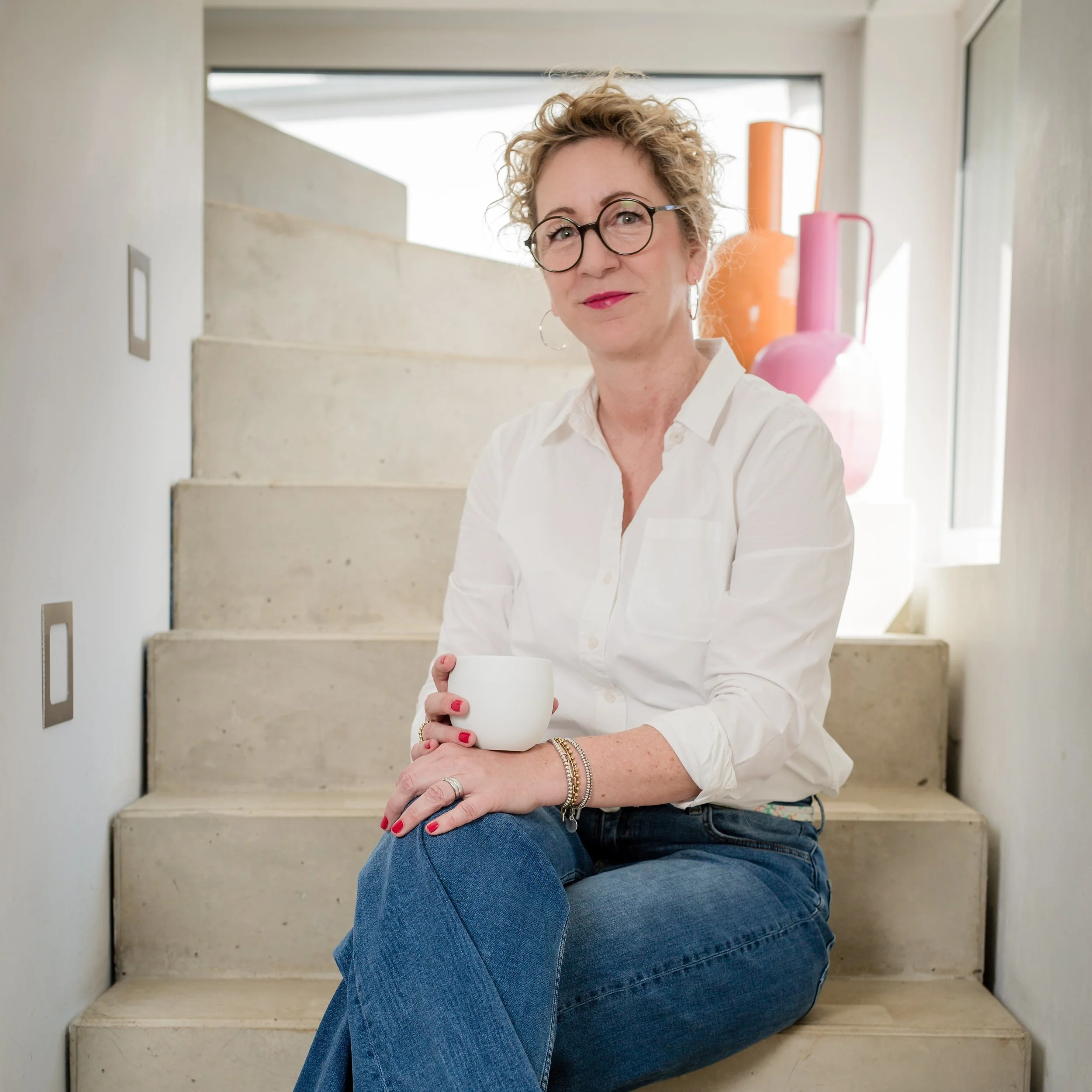 Frau mit lockigem Haar, Brille, weißen Hemd und blauen Jeans sitzt auf Stufen und hält eine weiße Tasse, im Hintergrund große Fenster